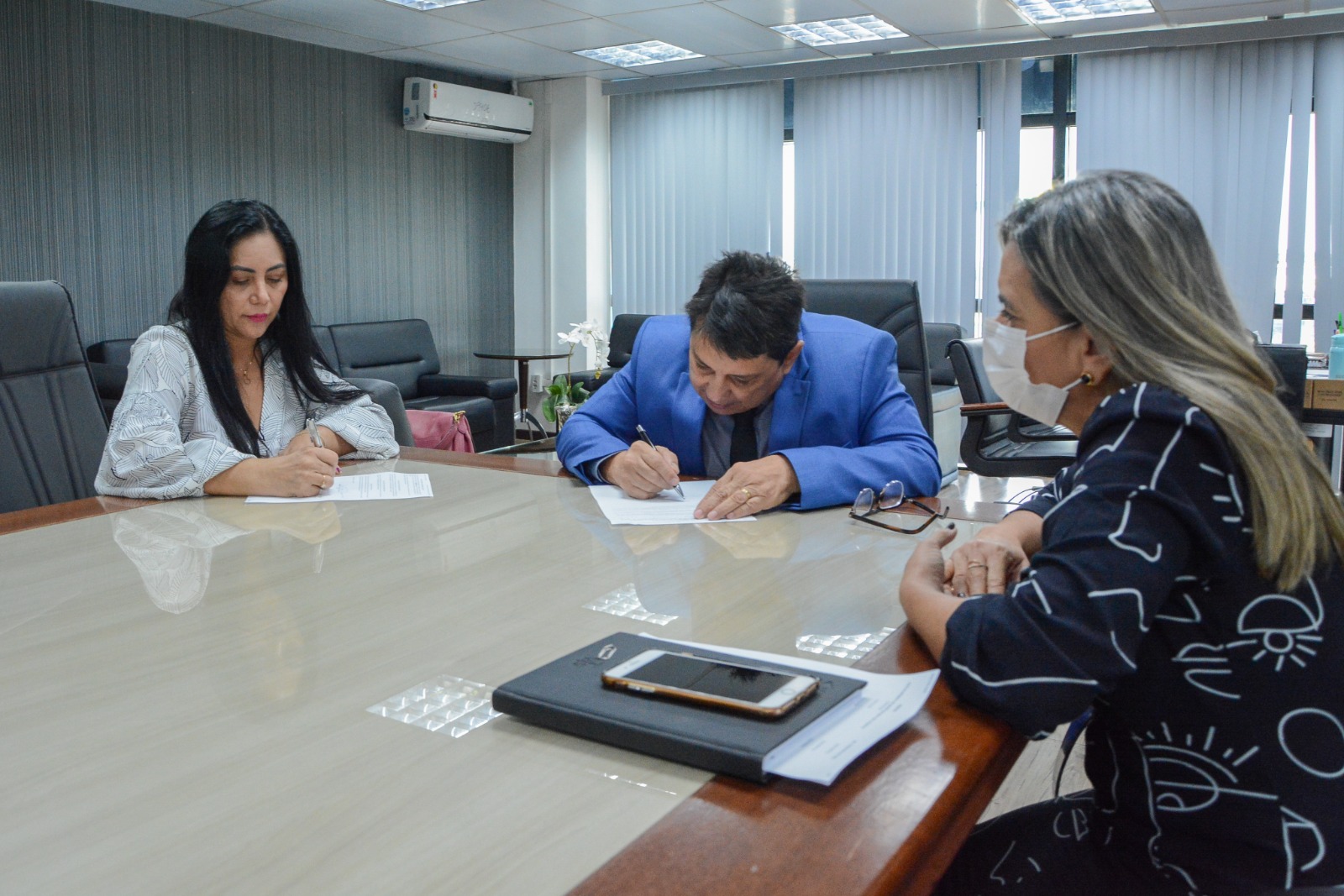 foto colorida do presidente do TJRR, desembargador Jésus Nascimento em uma mesa assisnando o termo de compromisso junto com duas servidoras da prefeitura