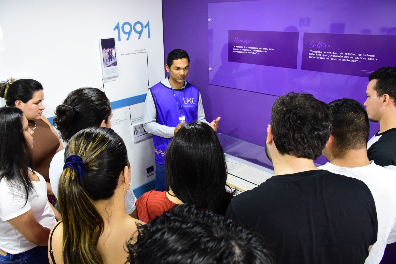  Imagem colorida mostra os alunos da UFRR de costa observando a explicação do historiador do Centro de Memória e Cultura do Poder Judiciário de Roraima, Hugo Mendes. 