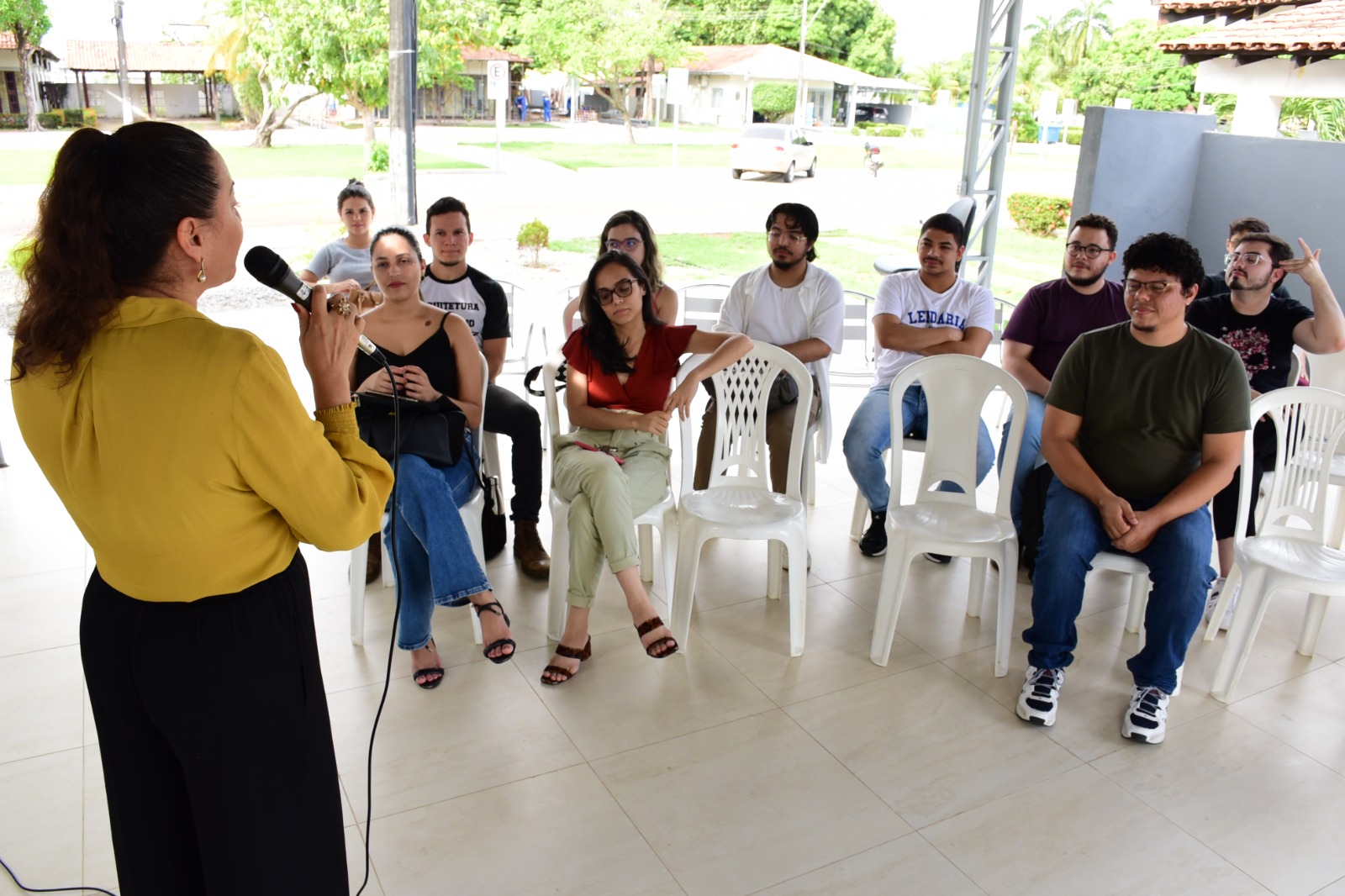 Imagem colorida mostra a coordenadora do Centro de Memória e Cultura, Olane Matos de costas, em pé e segurando o microfone na mão em frente a um grupo de Graduandos do curso de Arquitetura. Os estudantes estão sentados em cadeiras brancas olhando para a coordenadora.