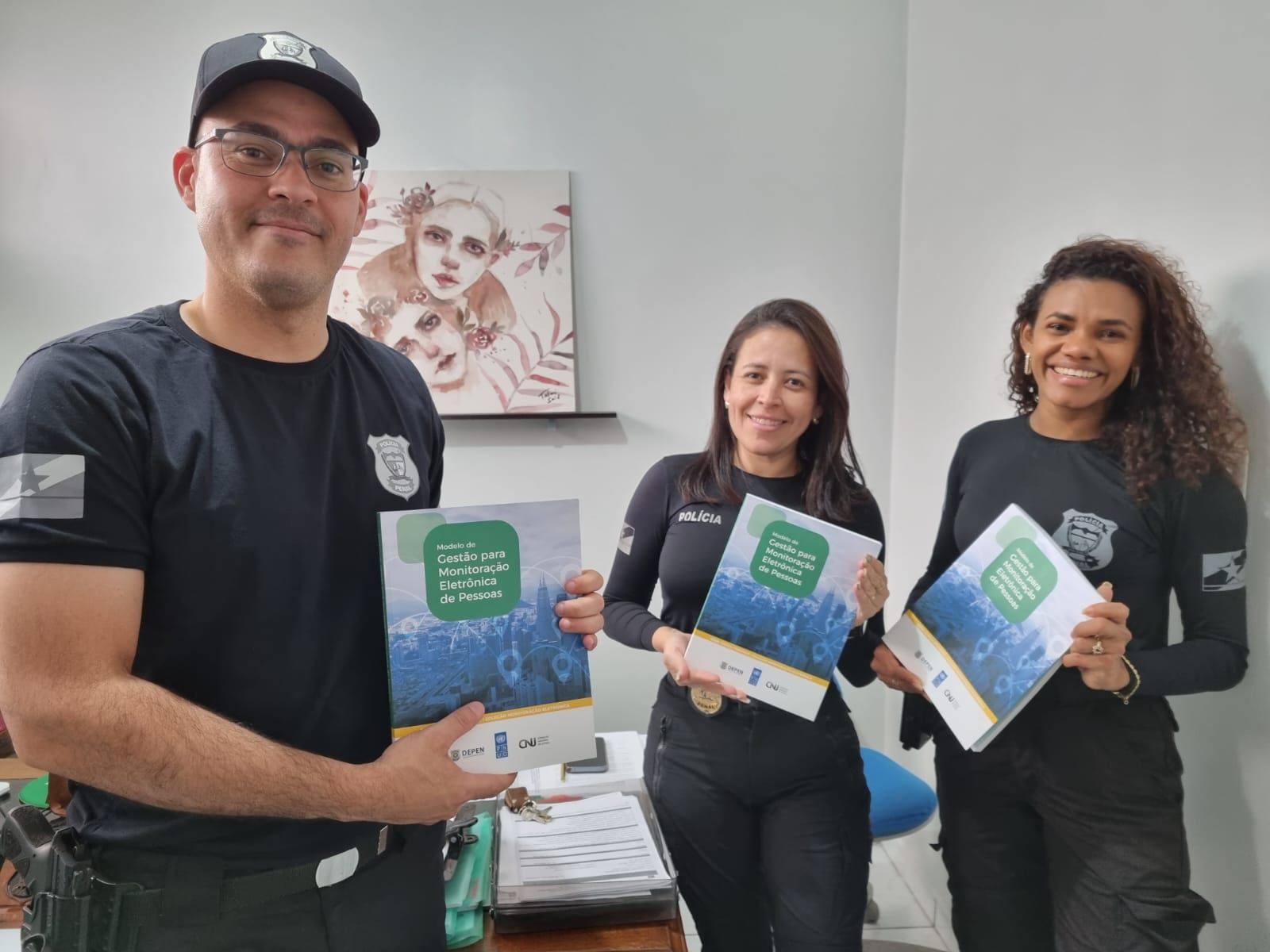 Imagem colorida mostra 3 agentes do Grupo de Monitoramento e Fiscalização do Sistema Carcerário (GMF), sendo estes 1 homem e duas mulheres em pé em uma sala com paredes brancas, segurando o manual de gestão para monitoração eletrônica de pessoas.