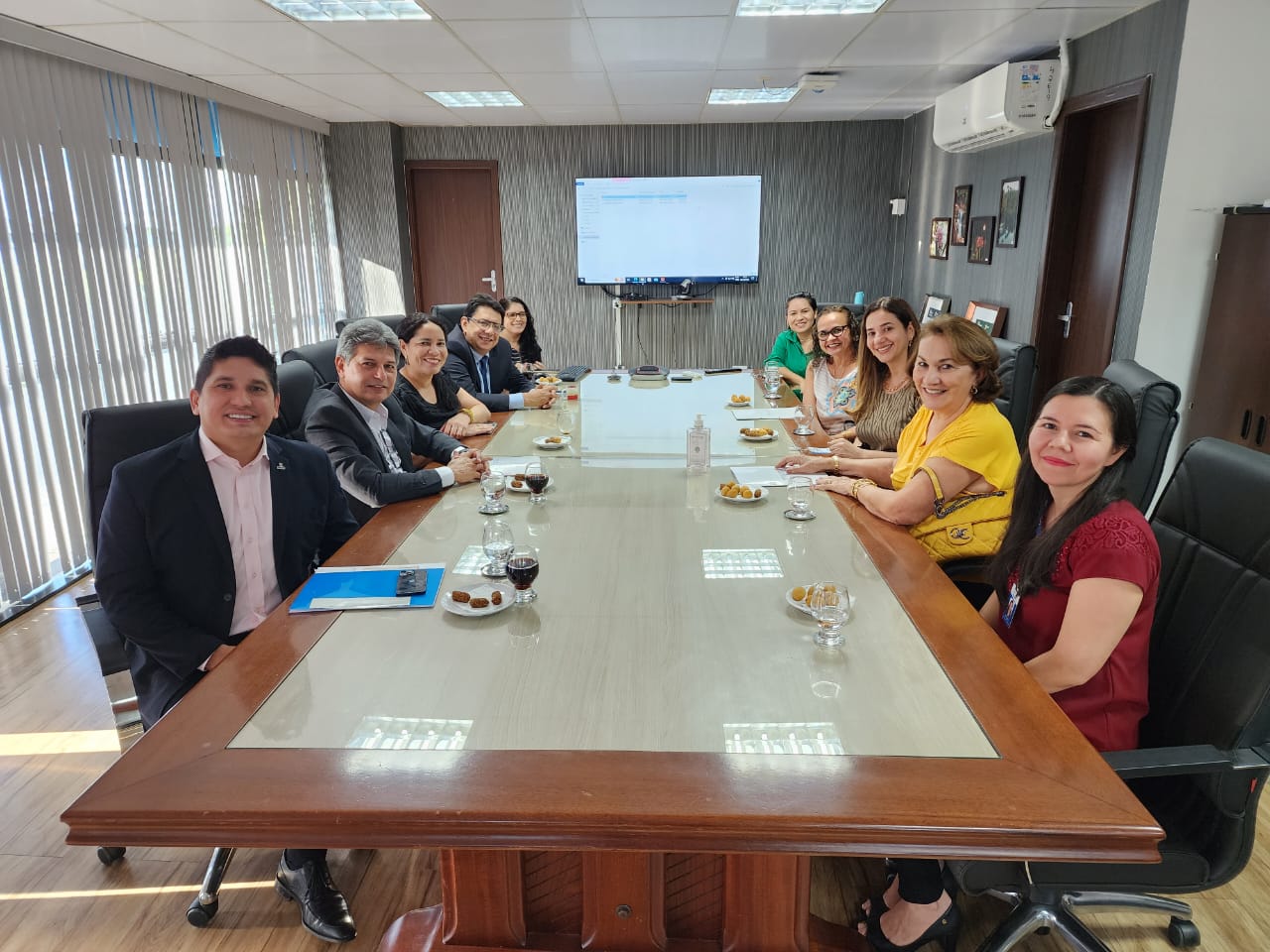 oto colorida mostra o presidente do TJRR, desembargador Jésus Nascimento, reunido na mesa de reunião da sala da presidência com os juízes titulares das Varas da Infância e Juventude, Parima Dias Veras e Marcelo Oliveira, a juíza titular da Vara da Justiça Itinerante, Graciete Sotto Mayor, a conselheira do TCE, Cilene Salomão e a defensora pública Emira Salomão. Além da secretária e a assessora executiva da Secretaria de Gestão de Magistrados, Fernanda Catanhede e Franciza Veríssimo,  a assessora Ana Moreira e a assessora executiva da Secretaria de Infraestrutura e Logística do Poder Judiciário de Roraima, Claudete Silva. Todos estão posando e sorrindo para a foto. Na sala também estão objetos como televisão e quadros artísticos. Abaixo está o texto “PROJETOS SOCIAIS. Presidente do TJRR reúne TCE, DPE e magistrados para debater revitalização de prédio público