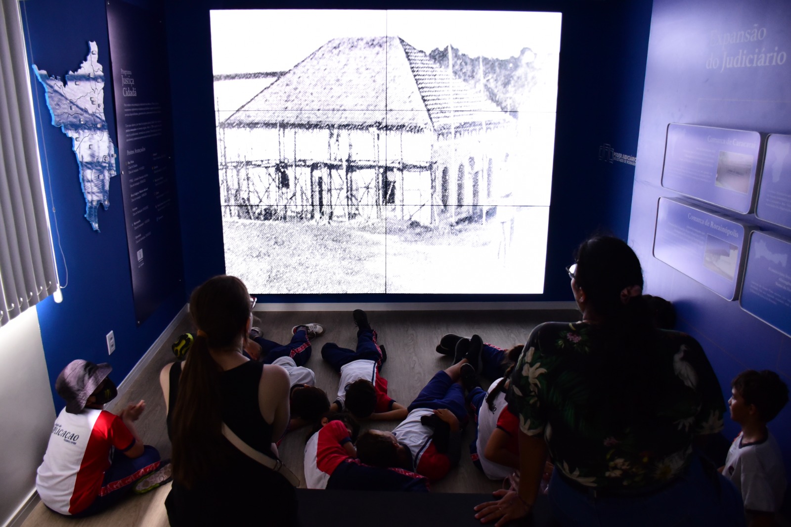 Foto colorida de alunos sentados no chão durante a exposição online do CMC