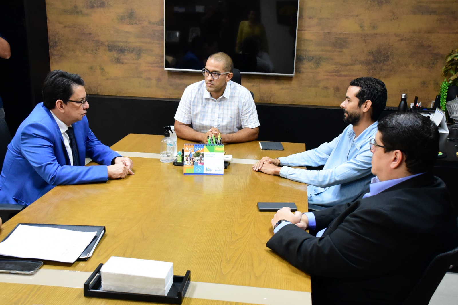 Imagem colorida mostra o presidente do Tribunal de Justiça de Roraima (TJRR), desembargador Jésus Nascimento, sentado na lateral de uma mesa marfim. Na ponta da mesa está  o prefeito de Boa Vista, Artur Henrique, do lado oposto, sentados, estão dois colaboradores observando a conversa.