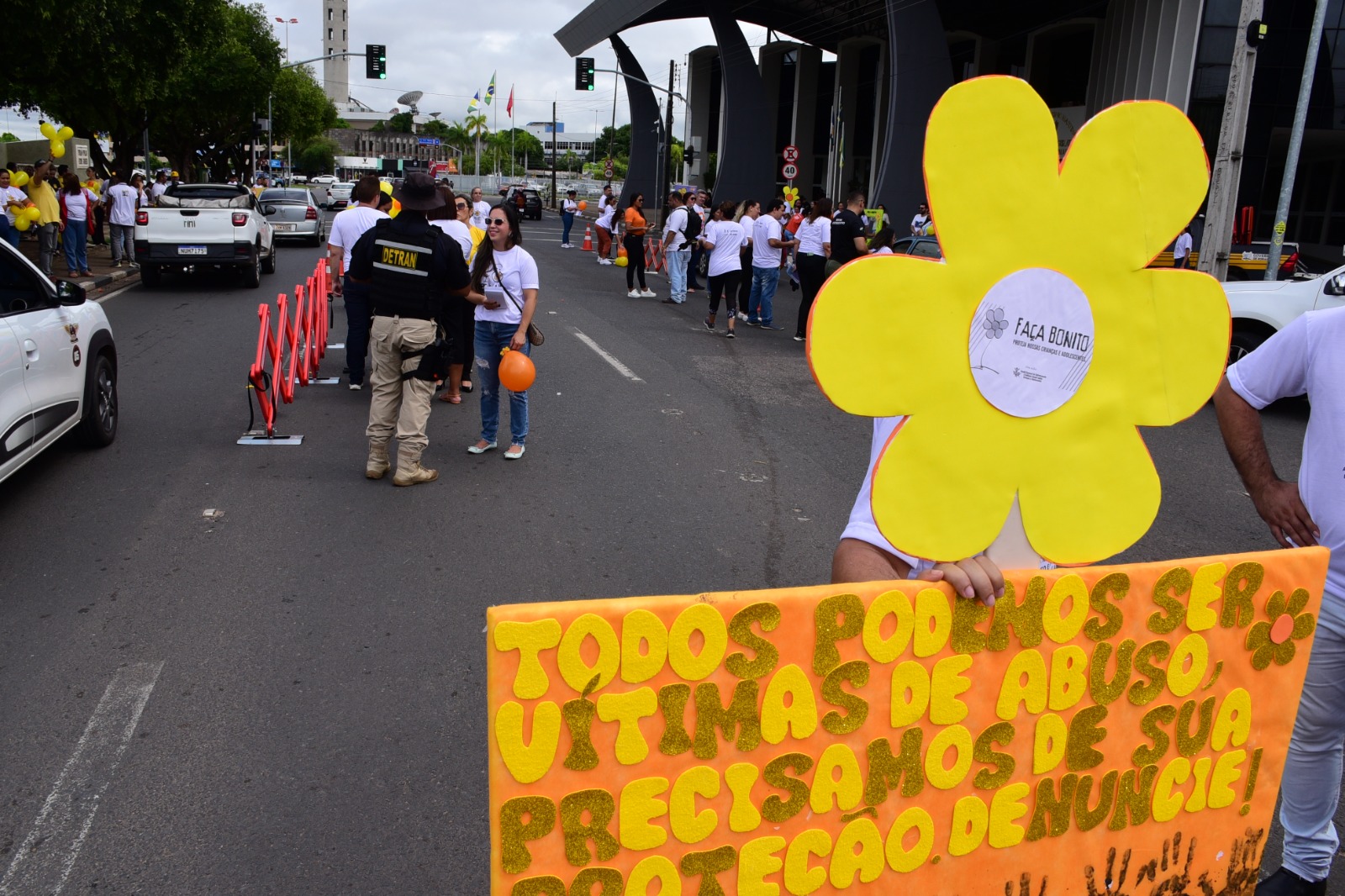 Imagem colorida mostra uma pessoa fantasiada de uma flor amarela segurando uma placa na cor laranja com a frase, “Todos podemos ser vítimas de abuso, precisamos de sua proteção, denuncie!”, nas cores amarela e dourada. Ao fundo há pessoas vestidas de branco segurando balões em uma via pavimentada.