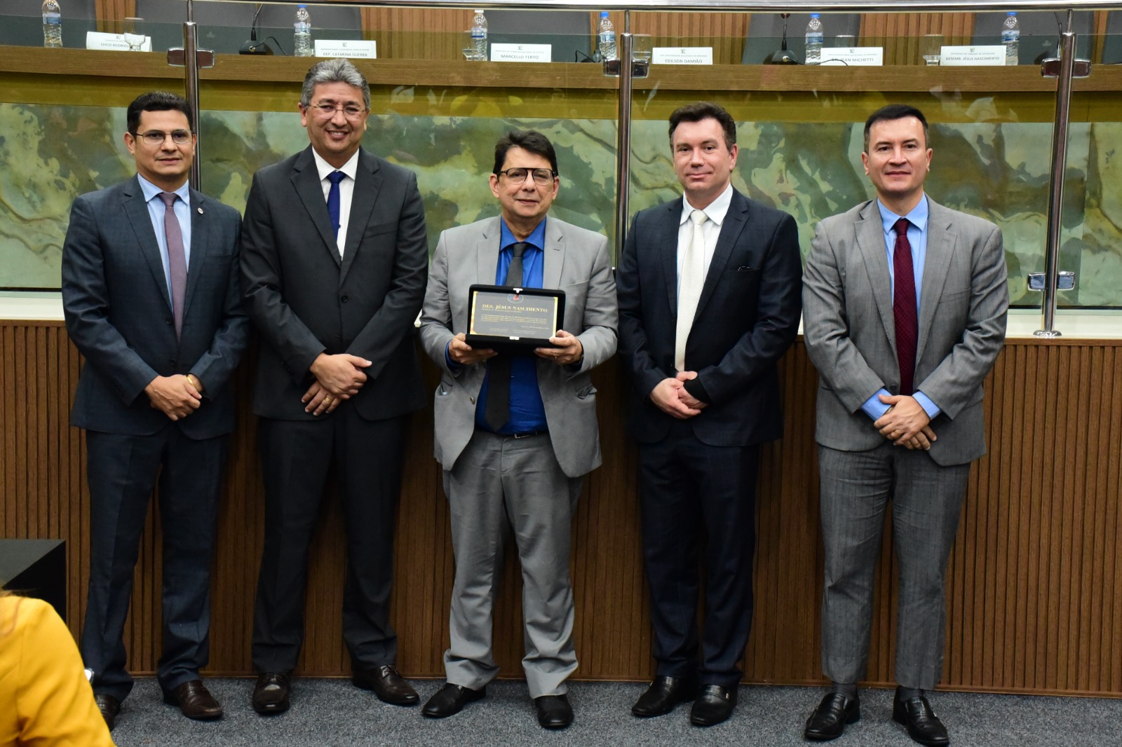 foto colorida do Presidente do TJRR, des Jésus Nascimento  segurando a placa de homenagame e pousando para foto ao lado de mais quatro autoridades