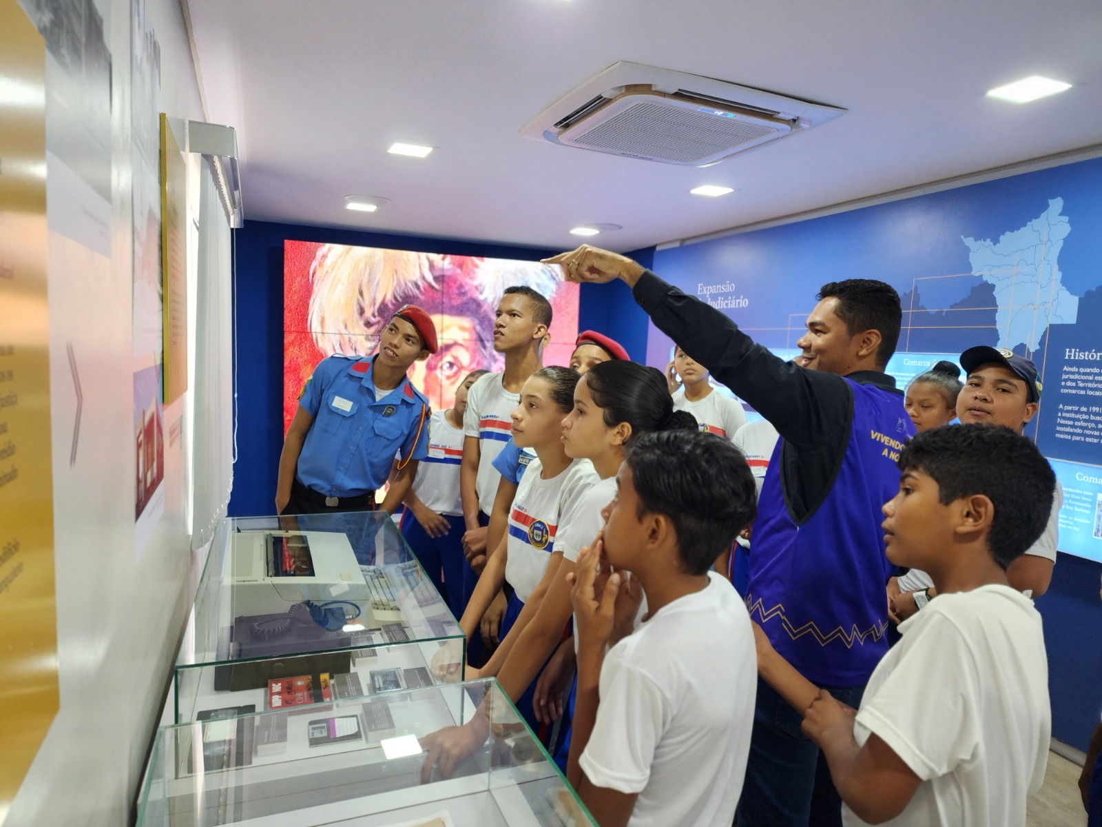  Foto mostra 12 alunos de costa, atentos à orientação do servidor do Centro de Memória e Cultura. Atrás dele, na parede, a foto mostra parte de uma linha do tempo do Tribunal de Justiça de Roraima.