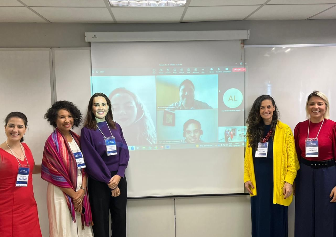 Imagem colorida mostra a coordenadora do NPI, Janaíne Voltolini, ao lado de outras quatro mulheres, em pé, posando para foto. Todas estão ao lado de um telão onde aparecem os participantes virtuais do evento.