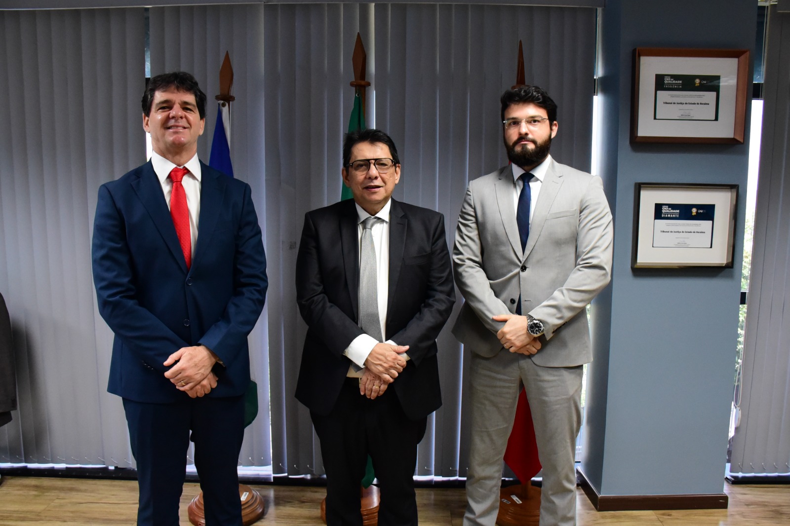 Foto colorida mostra o desembargador Jésus Nascimento, presidente do Tribunal de Justiça de Roraima, posando para foto com o superintendente da Polícia Federal, Ronaldo Guilherme Campos e o delegado federal Caio Luchine. Todos estão de pé, de mãos cruzadas em frente a três bandeiras na sala da presidência do TJRR