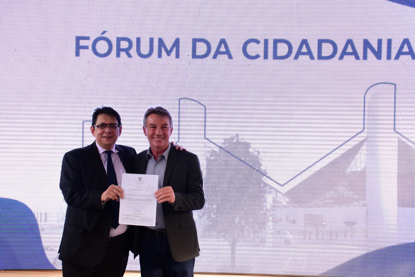 foto colorida do Presidente do TJRR, des Jésus Nascimento pousando para a  foto  junto com o governador Antonio Denario. Juntos estão segurando um contrato.