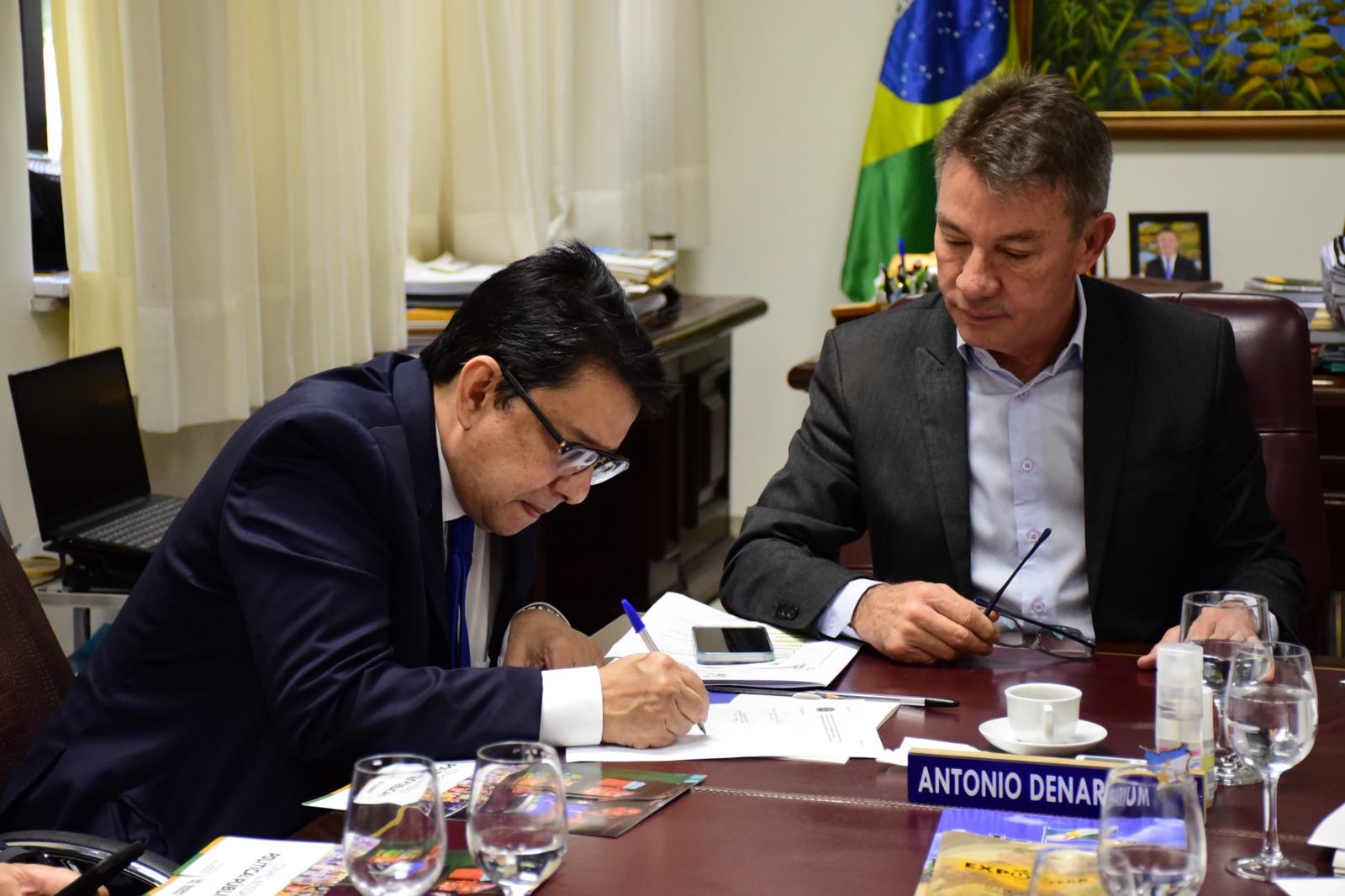 Foto colorida mostra o presidente do TJRR, desembargador Jésus Nascimento, e o prefeito o Governador do estado de Roraima, Antônio Denarium, assinando o termo de  regularização de imóvel. Ambos estão sentados na mesa. Atrás deles estão objetos, mobílias, quadros, a bandeira do Brasil e persianas. 