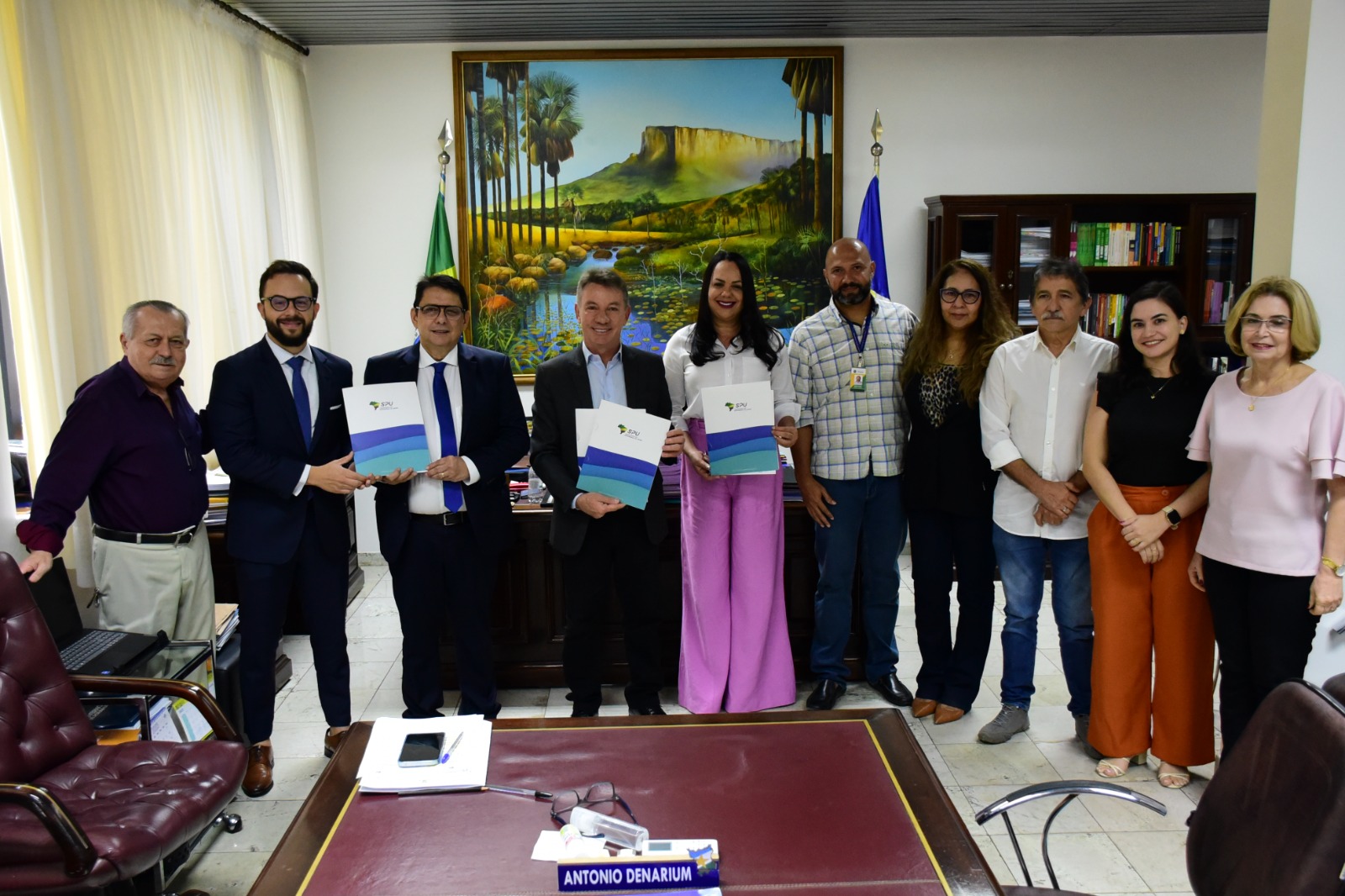  Foto colorida mostra o presidente do TJRR, desembargador Jésus Nascimento, sentado à mesa de reunião com o Governador do estado de Roraima, Antônio Denarium, o juiz auxiliar da presidência, Esdras Silva, juntamente com quatros servidoras e dois servidores das demais instituições, em pé, posando para foto. Atrás deles estão objetos, mobílias, um quadro ilustrando o monte Roraima, a bandeira do Brasil e do Estado e uma estante com livros. 