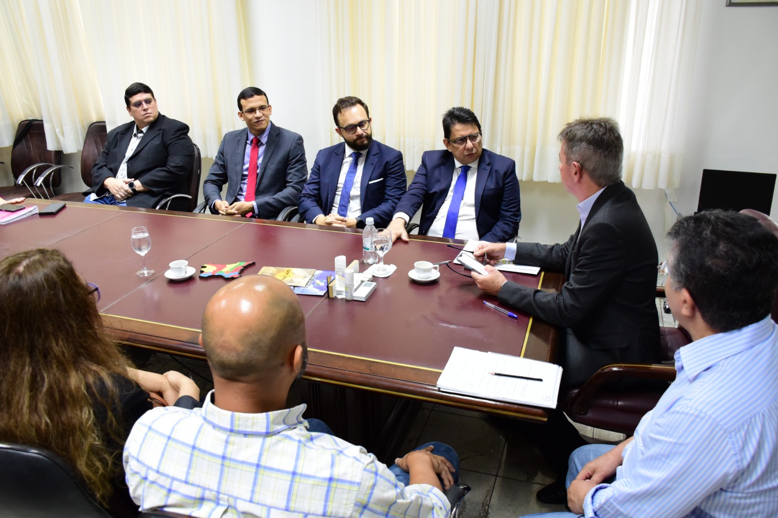  Foto colorida mostra o presidente do TJRR, desembargador Jésus Nascimento, sentado à mesa de reunião com o Governador do estado de Roraima, Antônio Denarium, o juiz auxiliar da presidência, Esdras Silva, o secretário geral do Judiciário, Henrique Tavares, juntamente com quatro  servidores das demais instituições, dos quais três estão de costas para foto. 