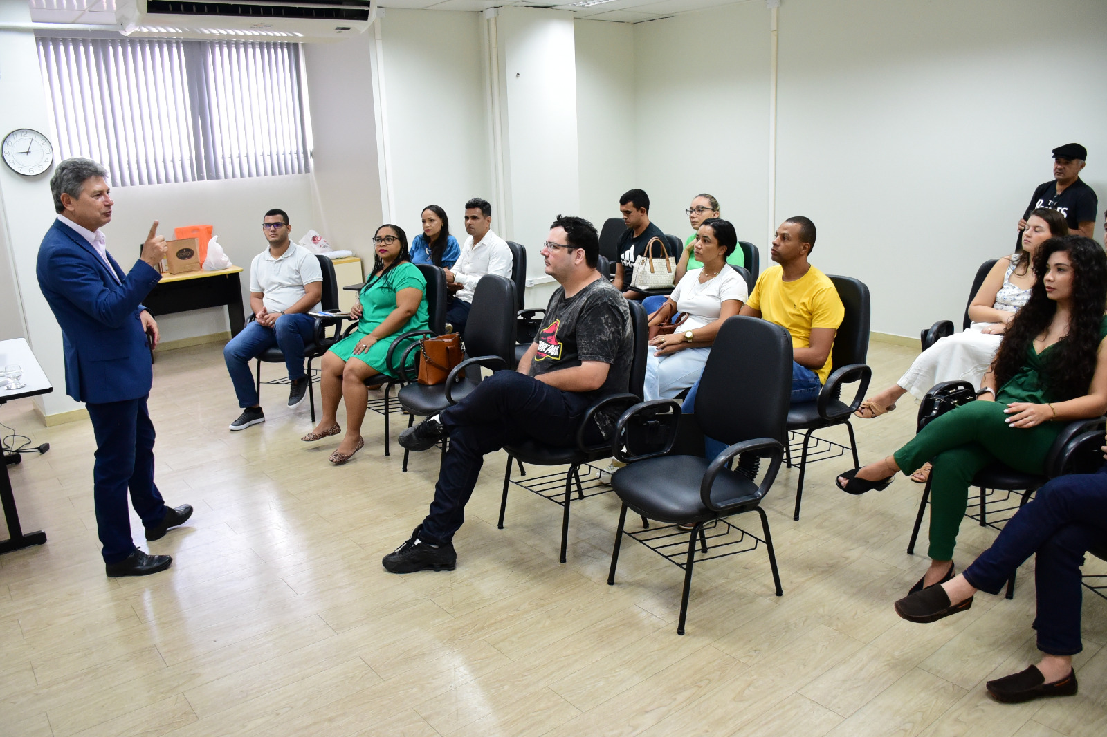 :Foto colorida mostra o Juiz titular da primeira Vara da infância e Juventude, Parima Dias Veras, em pé em um sala com paredes brancas ministrando para um grupo de homens e mulheres que estão assistindo a palestra, sentados em cadeiras pretas.
