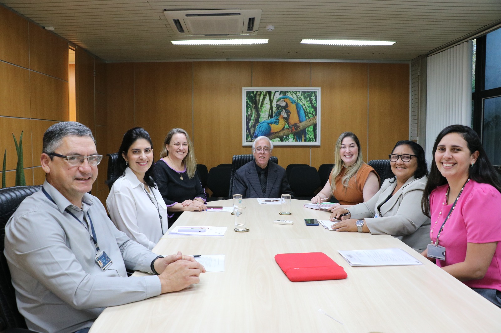 foto colorida mostra a juíza auxiliar da Corregedoria-Geral , Rafaella Holanda, e a subsecretária do setor de bens apreendidos do TJRR, Gicelda Assunção, sentadas em volta de uma mesa em formato retangular juntamente ao desembargador Fernando Mauro Moreira Marinho, Corregedor-Geral de Justiça de MS e presidente da Comissão de Alienação, a juíza auxiliar, Jacqueline Machado, e demais membros da Comissão de Alienação de Bens Apreendidos em Ações Penais do TJMS. Todos estão sorrindo para a foto, em uma sala com paredes cor madeira e um quadro com a imagem de duas araras. 