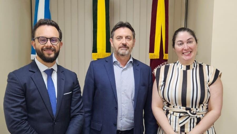 foto colorida mostra a direita o juiz auxiliar da presidência, o Procurador-Geral de Justiça do Ministério Público de Roraima, Fábio Bastos Stica e o Esdras Silva, a Juíza Auxiliar Interina da Corregedoria de Justiça, Rafaella Holanda. Todos estão em pé, posando para foto em frente a três bandeiras.