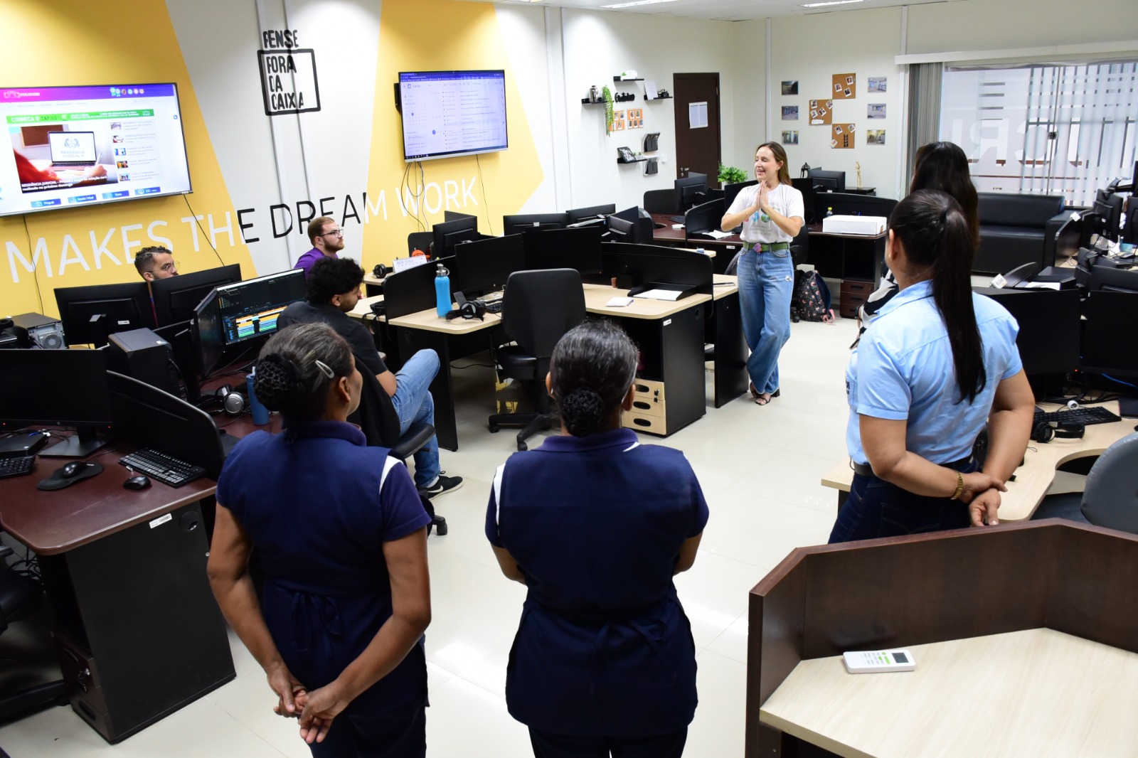 Foto mostra servidora do TJRR repassando orientações para outros servidores, que observam atentos, dentro de um espaço de trabalho onde existem estações e computadores.