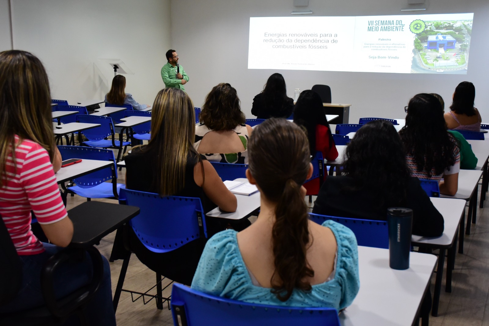 :Imagem colorida mostra cerca de 10 servidores do TJRR assistindo a palestra "Energias Renováveis e Alternativas para a Redução da Dependência de Combustíveis Fósseis", na sala da EJURR. A frente dos servidores está o palestrante em pé, segurando um microfone e olhando para as imagens projetadas na parede.