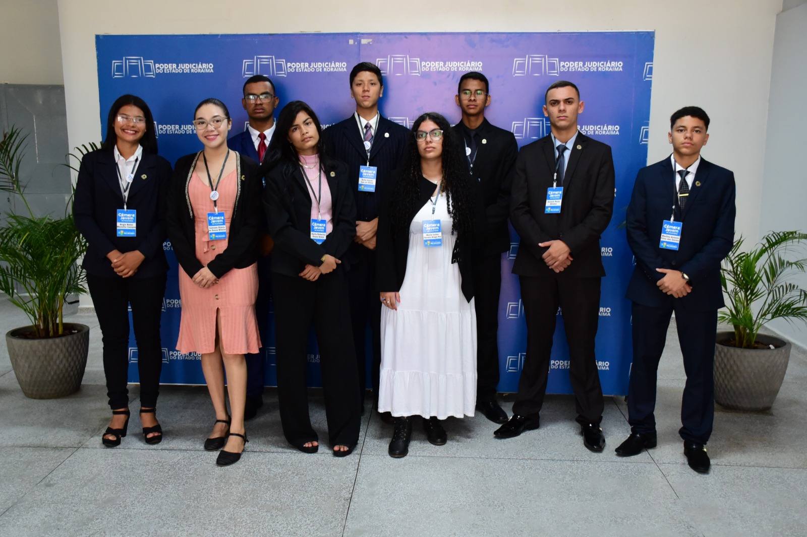 Foto colorida mostra nove estudantes do ensino médio de escolas públicas de Pacaraima, entre os quais quatro são mulheres. Todos estão de pé, identificados com crachás azuis, olhando para a câmera. Atrás dos alunos há um painel azul com diversos logotipos do Poder Judiciário. 