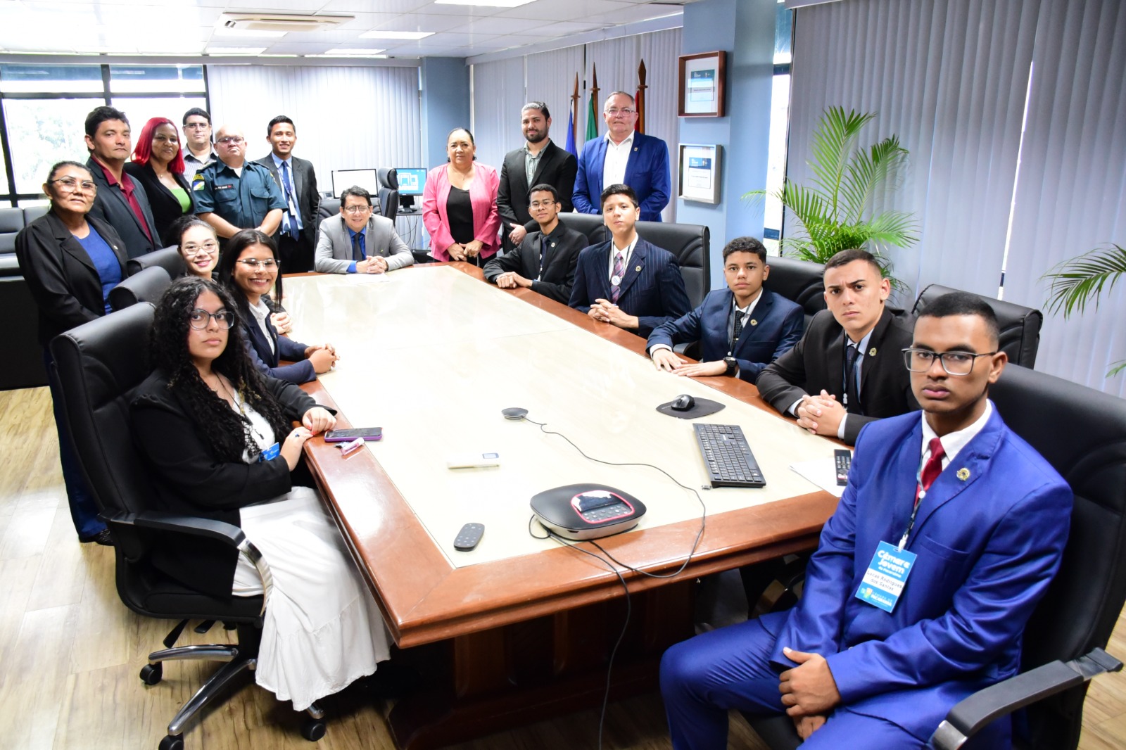 Foto colorida mostra o presidente do TJRR, desembargador Jésus Nascimento, sentado na ponta da mesa de reunião na sala da presidência do TJRR.  No lado esquerdo e direito do presidente, em pé,  estão os representantes dos poderes Judiciário, Executivo e Legislativo da Capital. Ao redor da mesa estão os nove alunos das escolas públicas de Pacaraima.