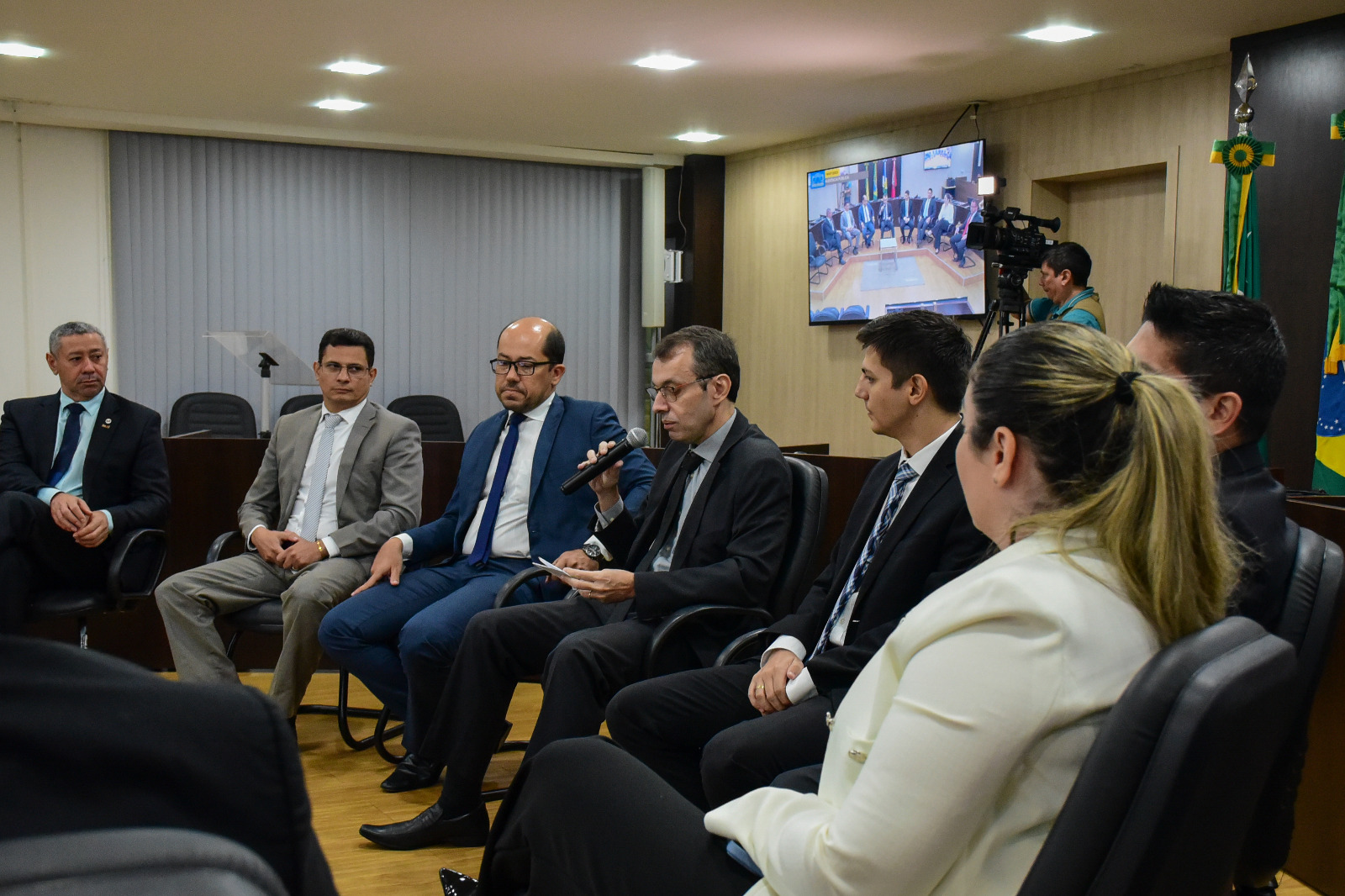 foto colorida mostra o desembargador Erick Linhares  falando ao microfone ao lado do juiz Rodrigo Delgado e do representante do MPRR. Demais autoridades que compõem o semi-círculo frente à tribuna do Tribunal Pleno acompanham a fala do desembargador. Ao fundo da imagem está uma TV fixada na parede e um cinegrafista com uma câmera.