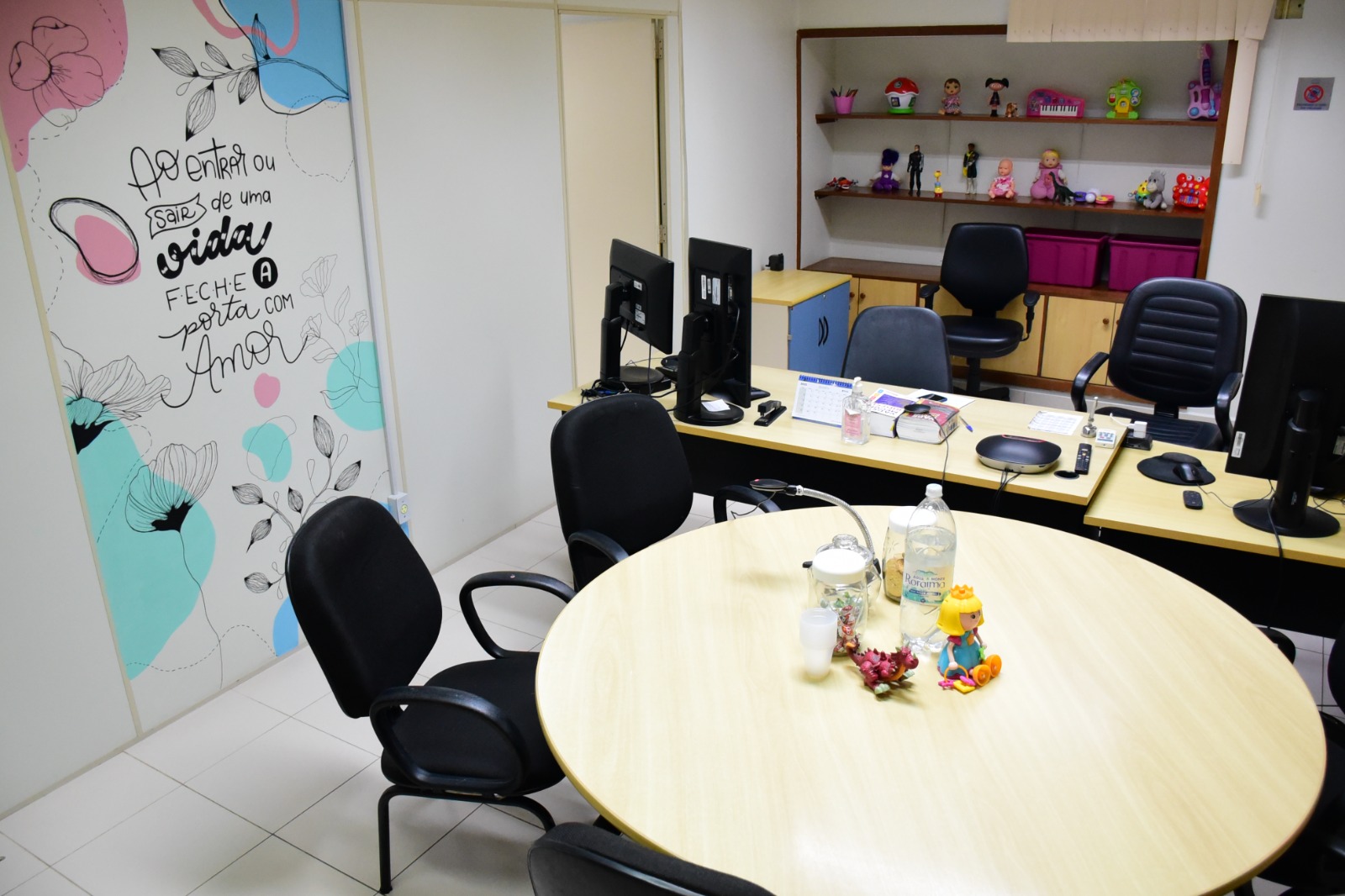 foto colorida de uma sala lúdica com flores e uma frase escrita e na parede de trás tem uma prateleira com bonecas 