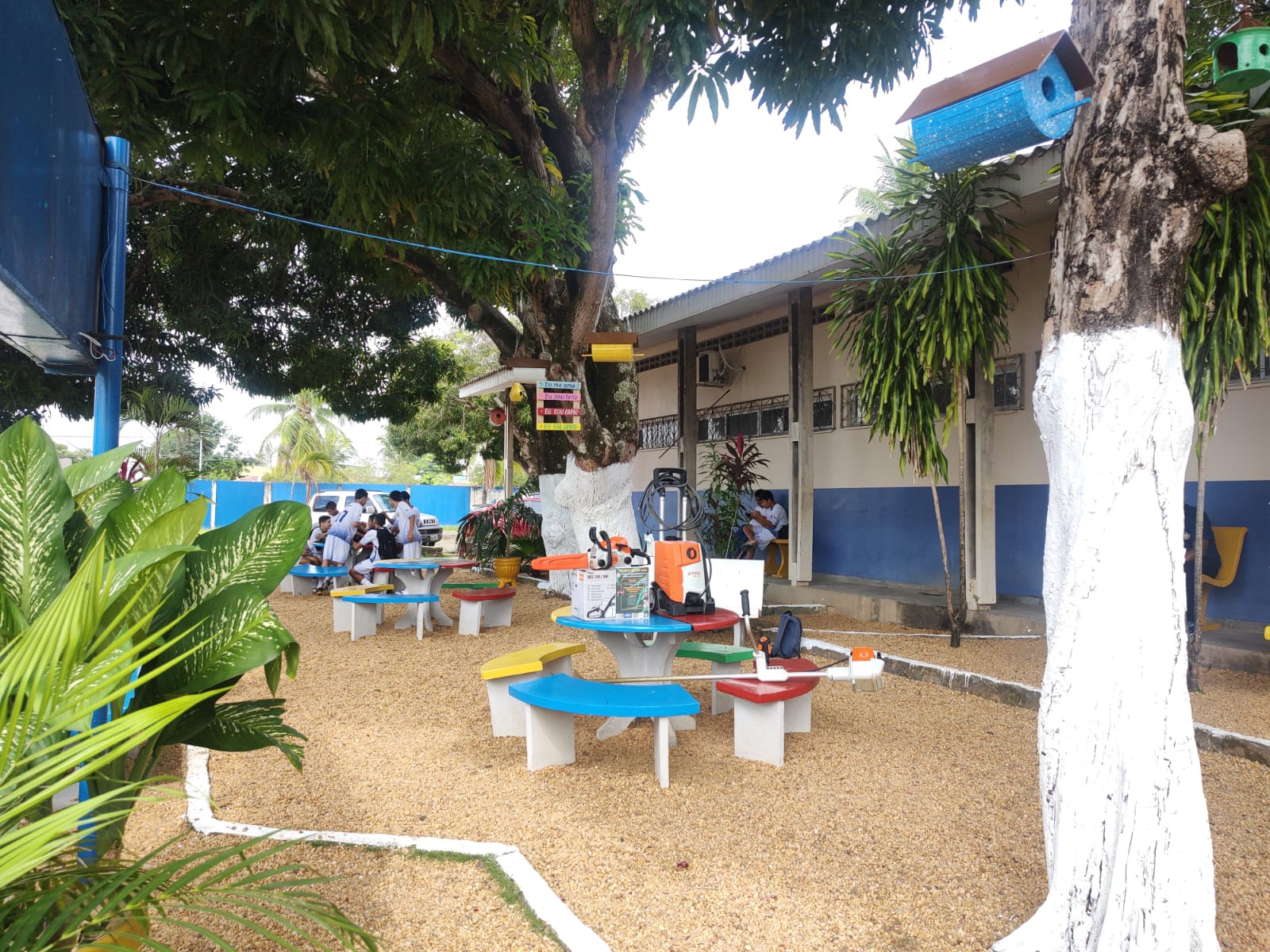  foto colorida em plano aberto mostrando o projeto feito no jardim e alunos sentados bem ao fundo nas mesas, no centro da foto está a mesas e bancos de cimento coloridas em azul, amarelo e vermelho, no canto da imagem está uma árvore com o tronco pintado de branco com casinha de passarinho pendurada.  Abaixo está o texto “VEPEMA Recursos destinados a projetos sociais são aplicados em ação ambiental de escola”