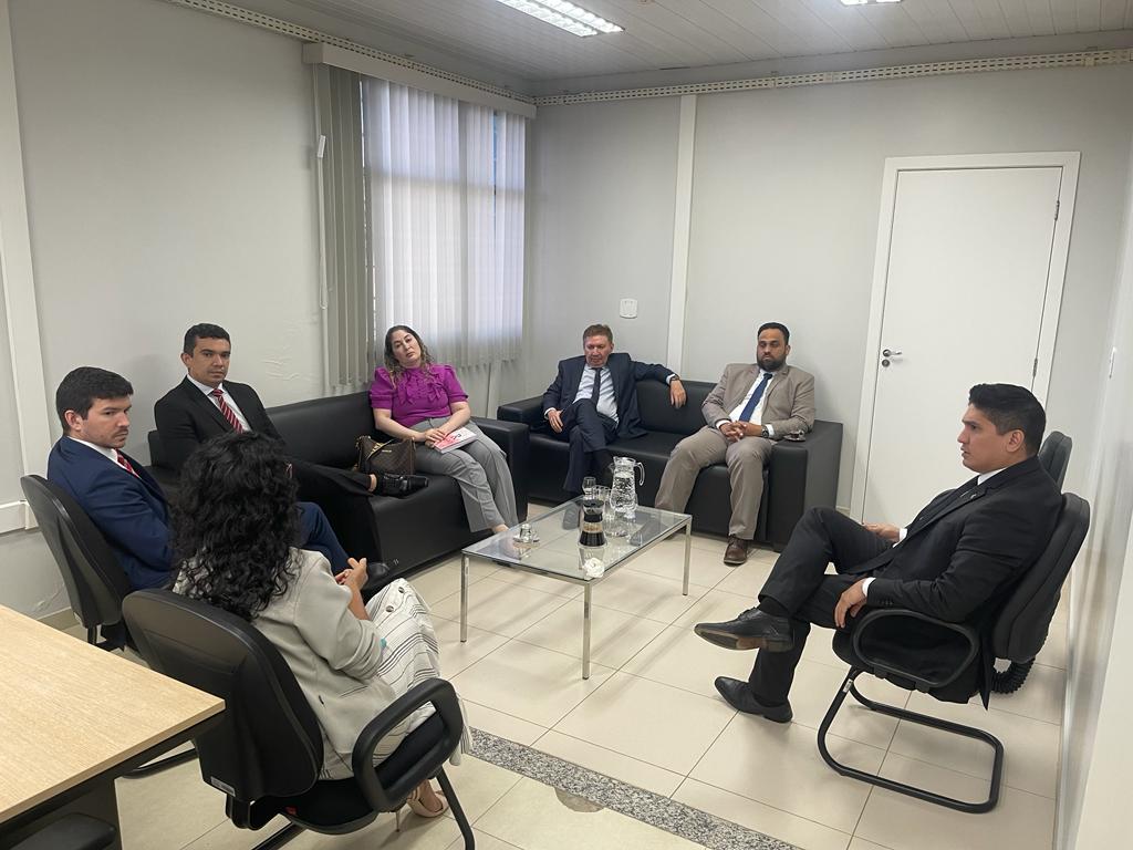 Foto colorida mostra o vice-presidente do TJRR, desembargador Ricardo Oliveira, o desembargador Almiro Padilha, corregedor-geral da Justiça do TJRR, desembargador Mozarildo Cavalcanti e o Corregedor Geral da Justiça do Piauí, desembargador Olímpio José Passos Galvão, em pé, posando para a foto. No fundo há 3 bandeiras hasteadas e uma cortina de persiana cinza cobrindo toda a parede de vidro.