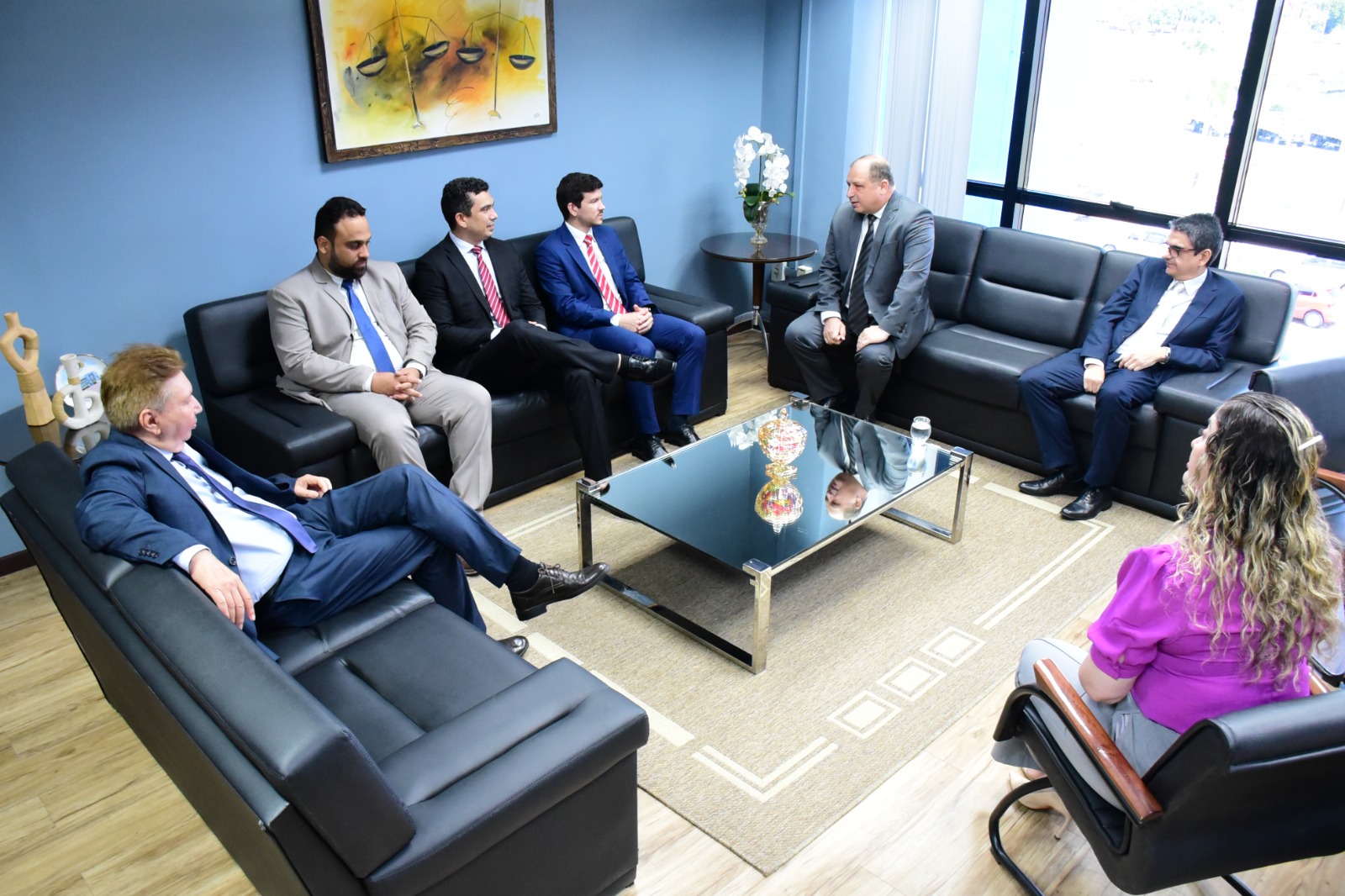  Foto Colorida mostra o vice-presidente do TJRR, desembargador Ricardo Oliveira, o desembargador Almiro Padilha, corregedor-geral da Justiça do TJRR, desembargador Mozarildo Cavalcanti, a juíza auxiliar da corregedoria, Rafaella Holanda, o Corregedor Geral da Justiça do Piauí, desembargador Olímpio José Passos Galvão, e os servidores do TJPI Elias Ribeiro, superintendente da secretaria da Corregedoria, Leandro Rodrigues, analista judicial do Núcleo de Aceleração de Projetos, Sávio Mota, o analista de sistemas, sentados em sofás pretos que formam um círculo ao redor de uma mesa de centro de vidro. No fundo, uma parede azul com um quadro pintado a mão com um desenho de duas balanças.