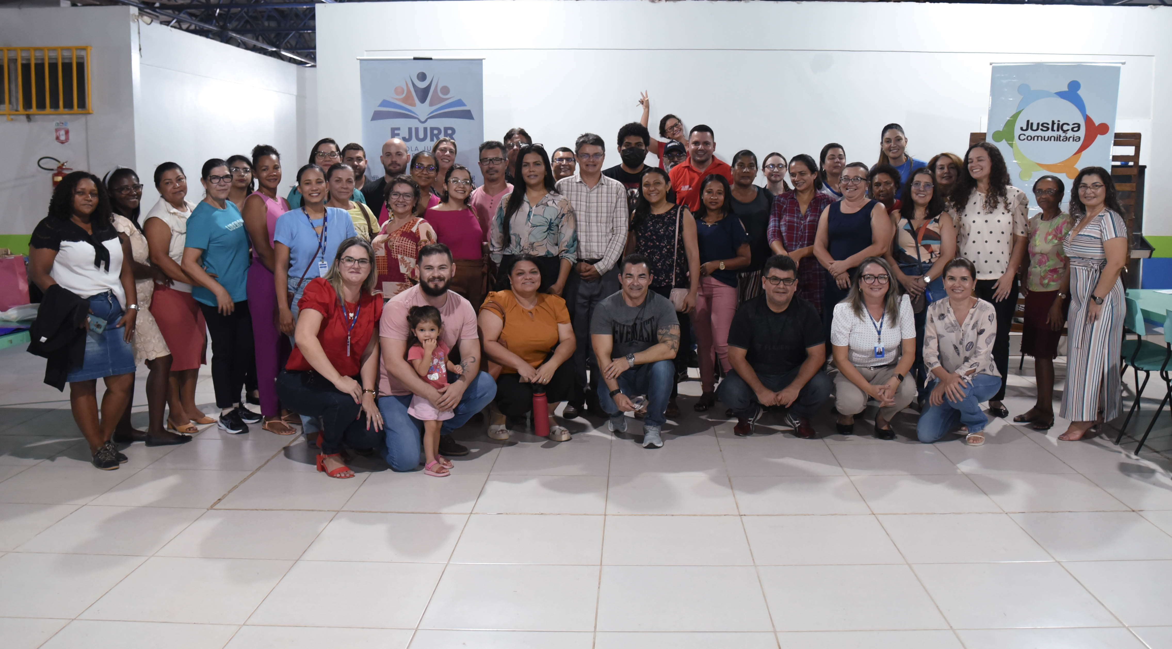 Pessoas reunidas em frente a um fundo branco com um banner do programa Justiça Comunitária e outro da EJURR
