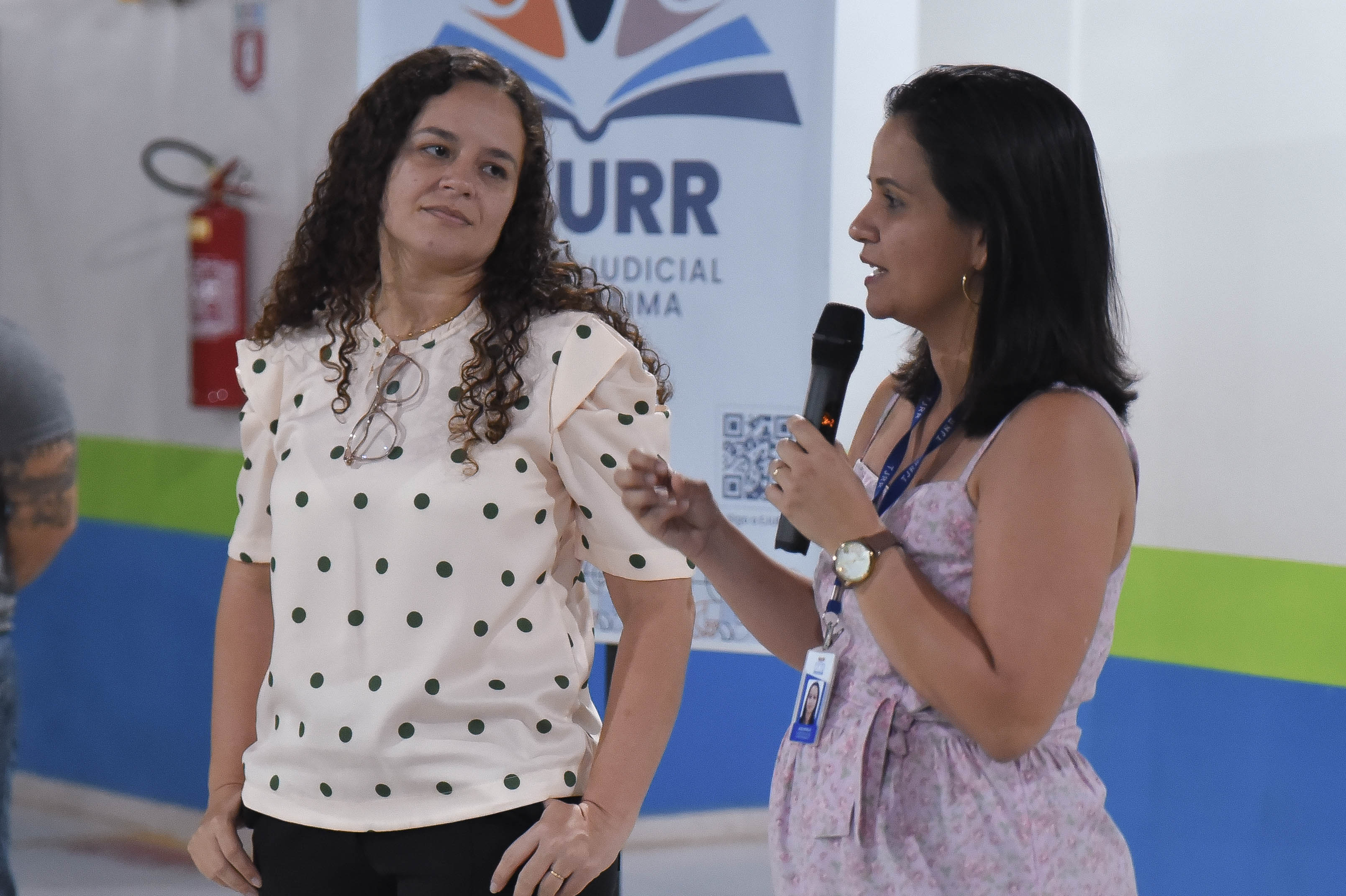 Uma mulher fala segurando um microfone enquanto outra observa. As duas estão em pé e em frente a um banner da EJURR.