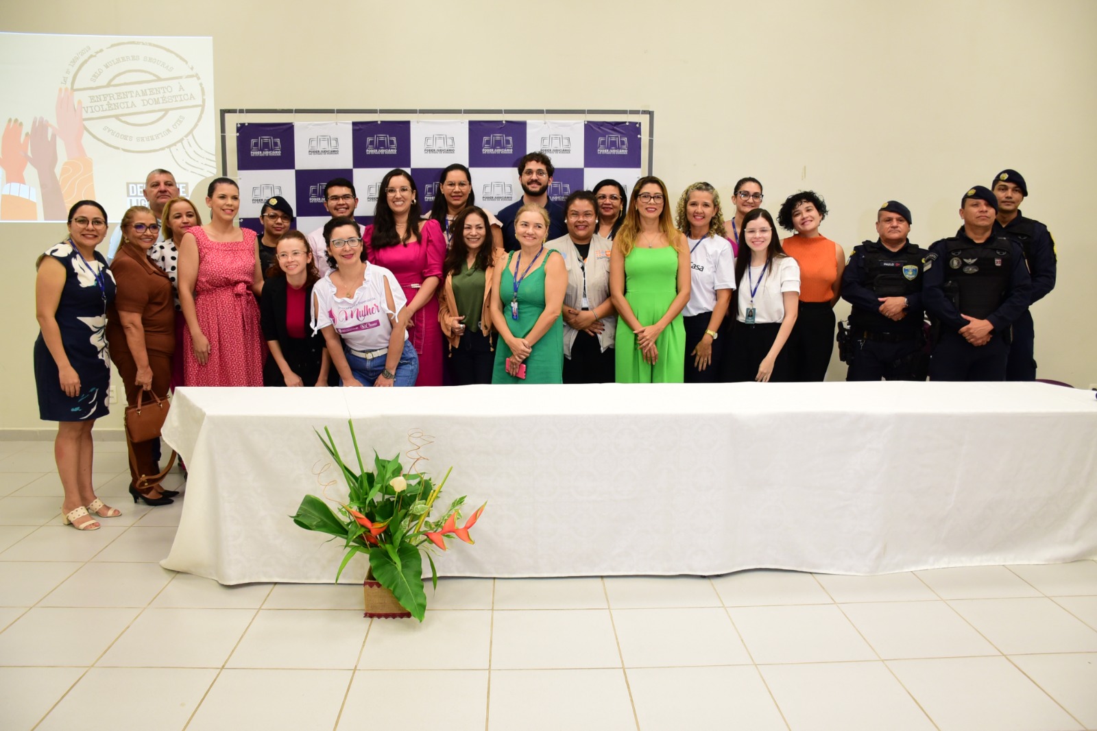  foto colorida mostra grupo de 24 pessoas posando para fotografia atrás de mesa branca. Na imagem está a juíza titular da Cevid, Suelen Alves, a chefe do Setor de Enfrentamento a Violência Doméstica do TJRR, Aurilene Mesquita, e mais 22 pessoas que participaram do lançamento do selo, entre servidores do TJRR, policiais fardados e convidados.