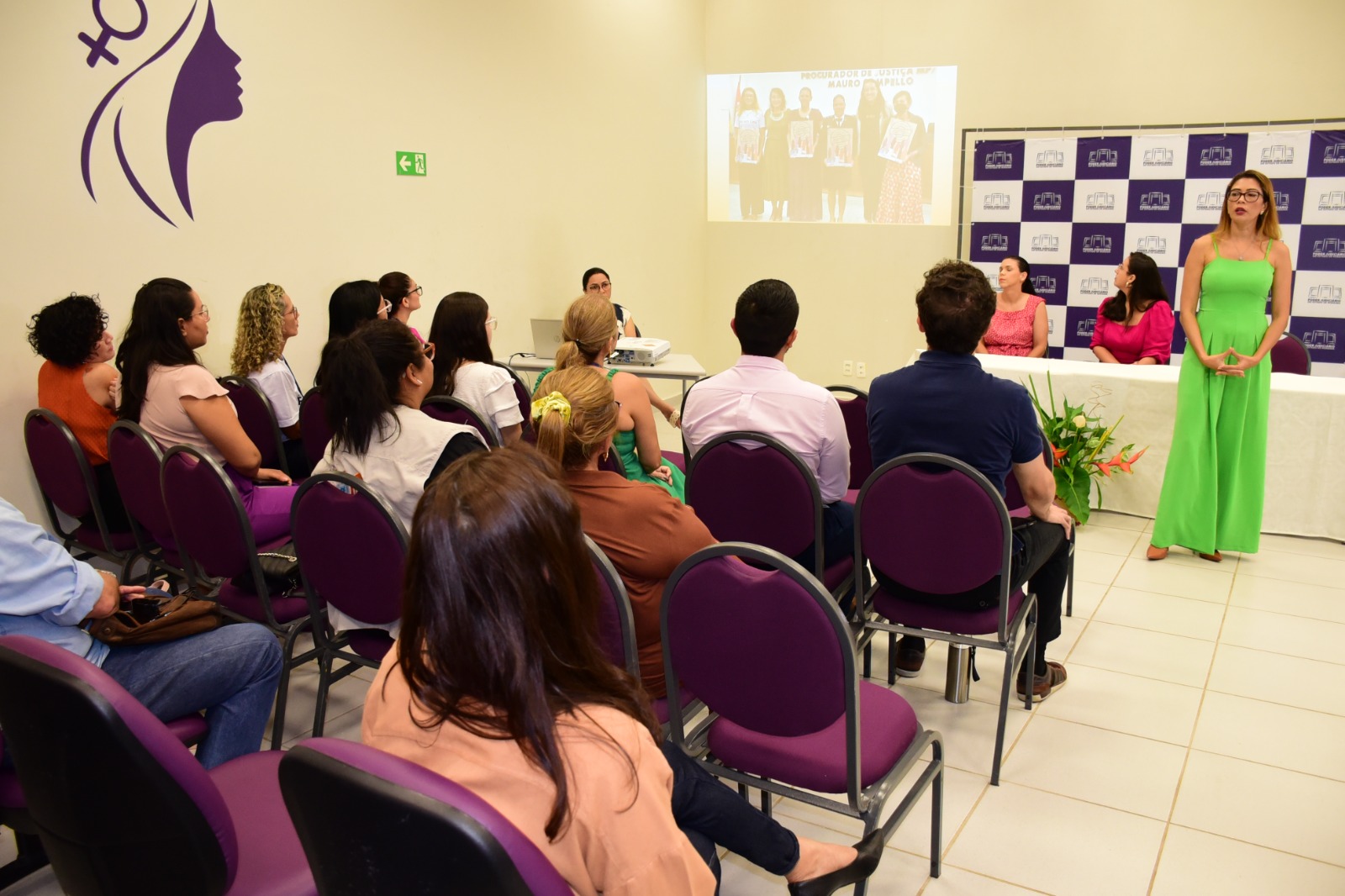  foto colorida mostra um grupo de pessoas sentadas assistindo apresentação do lançamento do Selo Mulheres Seguras. Na imagem está a chefe do Setor de Enfrentamento a Violência Doméstica do TJRR, Aurilene Mesquita palestrando para as pessoas durante o lançamento do selo e atrás dela encontram se a juíza titular da Cevid, Suelen Alves e a ganhadora do último Selo Mulheres Seguras, Nannibia Cabral, sentadas na mesa de autoridades.