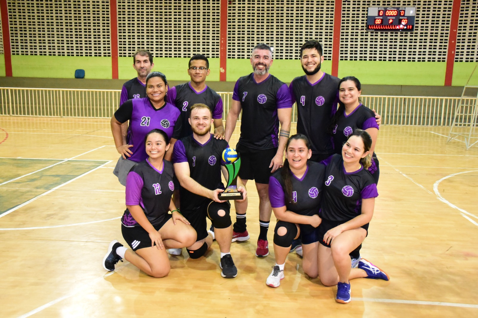 Foto colorida mostra as equipes de jogadores uniformizados, posando para a foto, estando três deles em pé no pódio segurando um troféu na mão.  Ao fundo uma parede verde com uma corrente de balões nas cores verde, amarelo, branco e azul