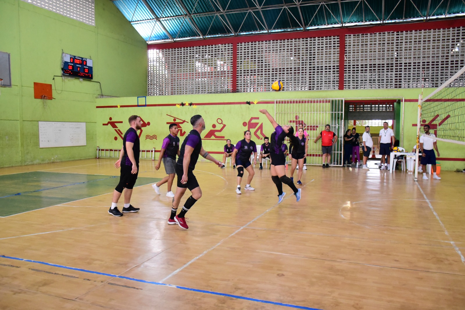  Foto colorida mostra uma partida de vôlei em uma quadra de esporte com paredes verdes