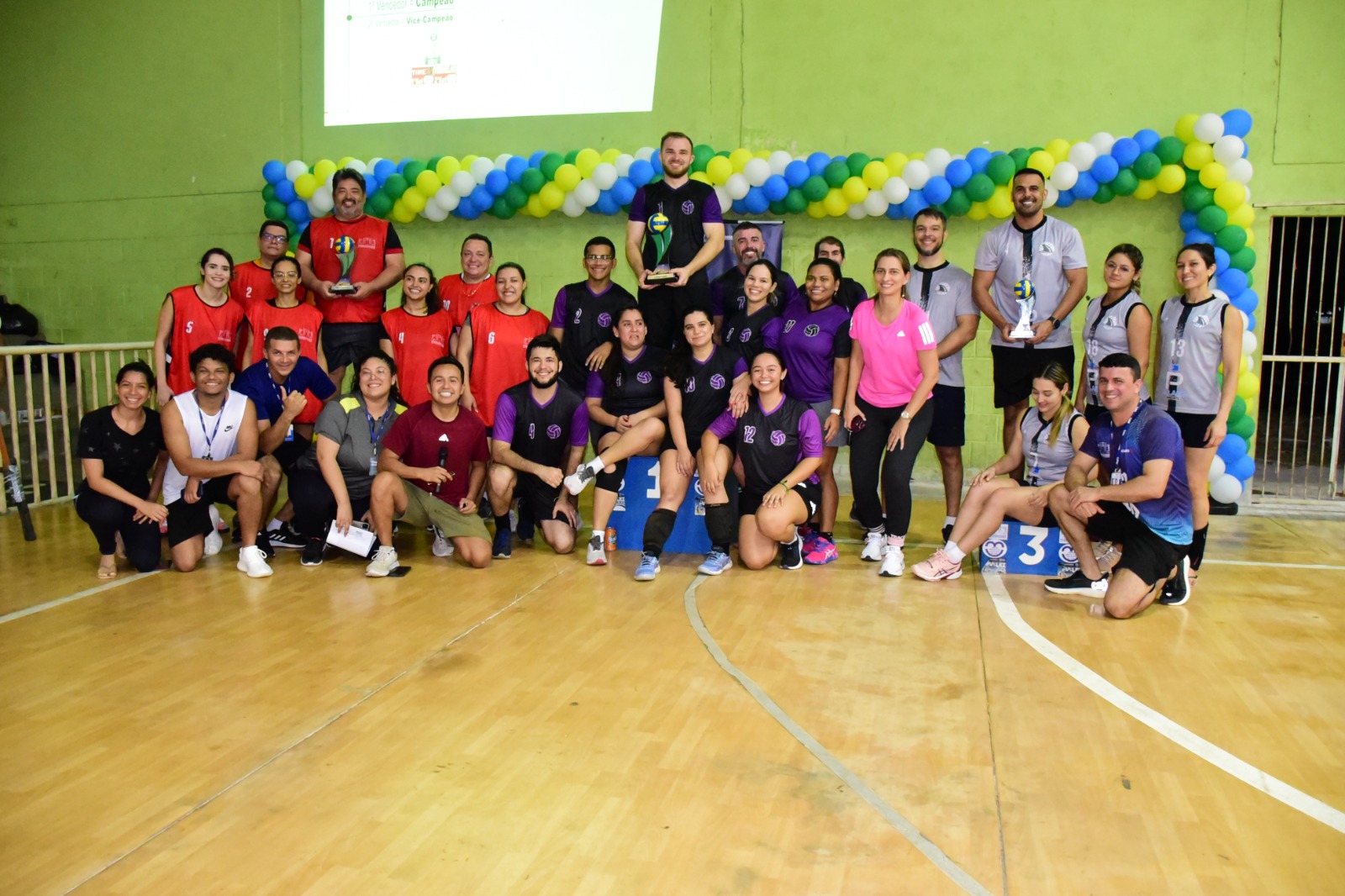 Foto colorida mostra as equipes de jogadores uniformizados, posando para a foto, estando três deles em pé no pódio segurando um troféu na mão.  Ao fundo uma parede verde com uma corrente de balões nas cores verde, amarelo, branco e azul