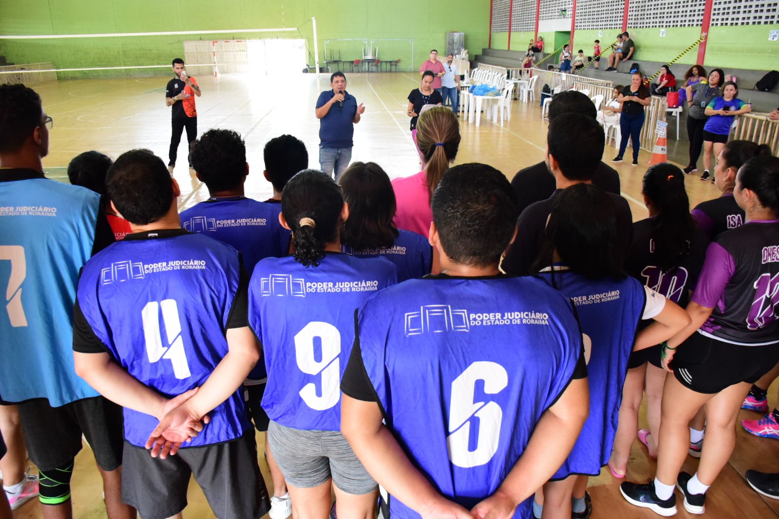  Foto colorida mostra uma equipe de jogadores vestidos com um uniforme preto com detalhes roxos, posando para a foto em uma quadra de esporte com paredes verdes. No centro, um dos jogadores  está segurando o troféu na mão