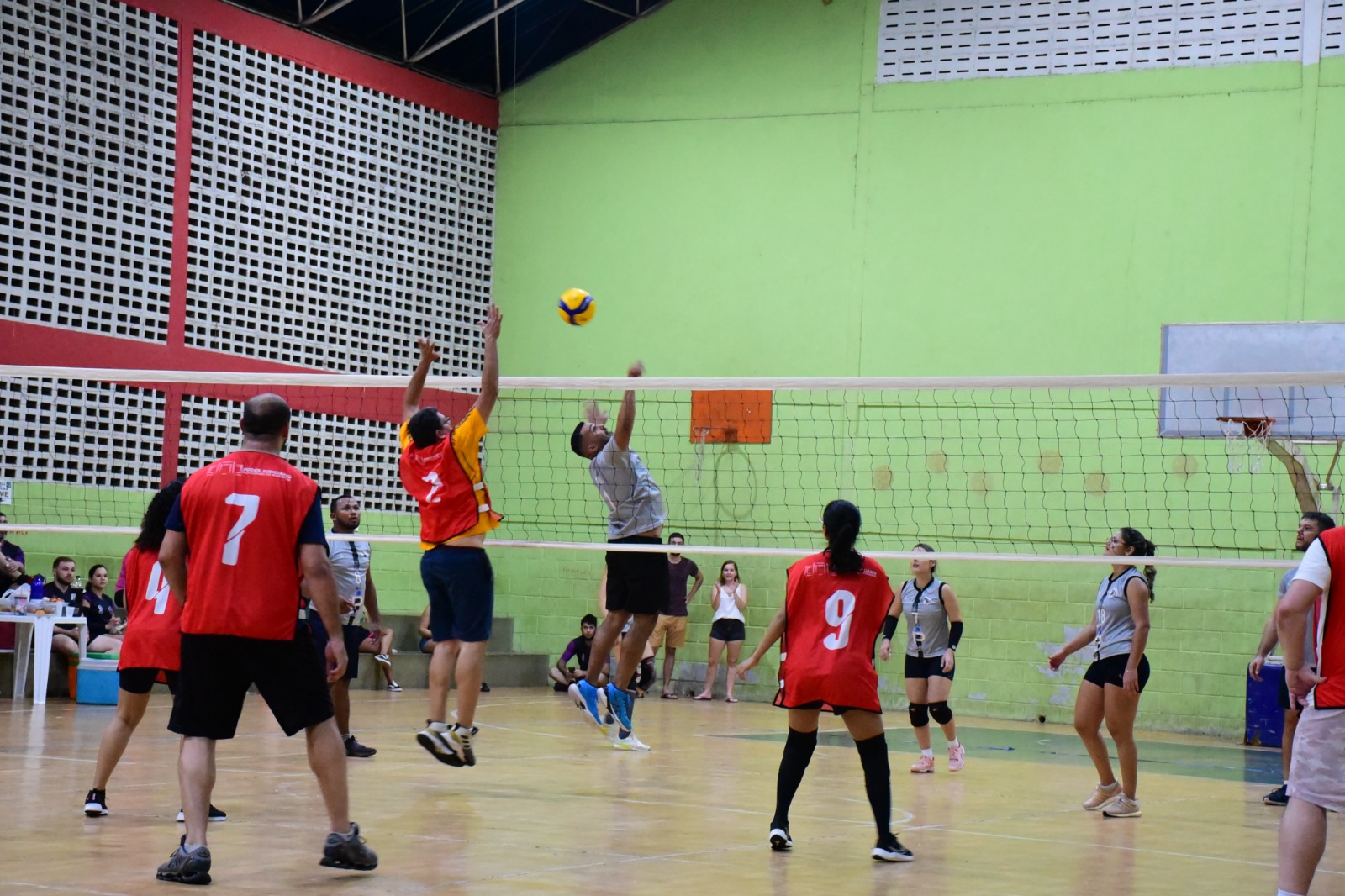 Foto colorida mostra uma partida de vôlei em uma quadra de esporte com paredes verdes