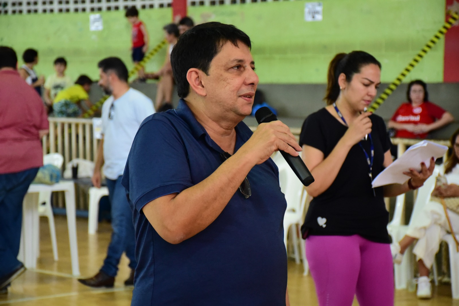  Foto colorida mostra o desembargador Jésus Nascimento com um microfone na mão em uma quadra de esporte com paredes verdes, discursando para servidores, que estão de costas para a câmera, vestidos de uniforme esportivo