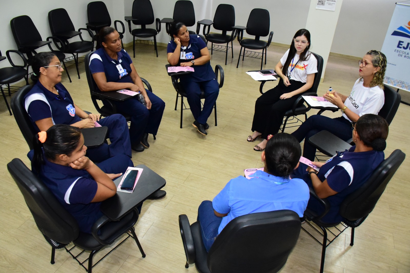 imagem colorida com um grupo pequeno de mulheres, servidoras terceirizadas do TJRR, sentadas em uma roda segurando ou encima da mesa da cadeira uma cartilha, rosa cartilha que exemplifica cada um dos temas debatidos durante a roda de convers