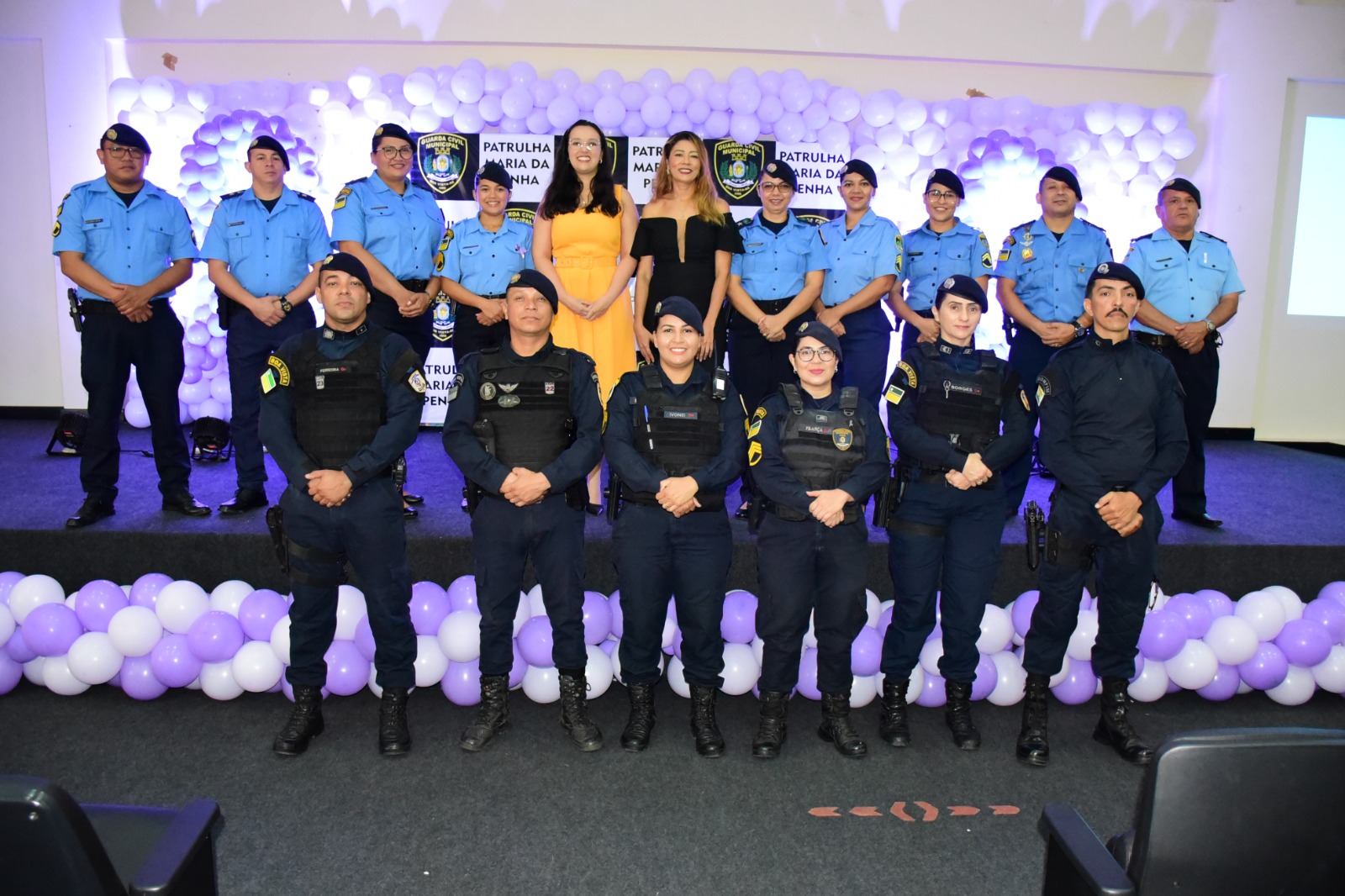 : imagem colorida com 16 Guardas Civis Municipais (GCM) fardados e alinhados de frente posando para foto. Ao centro está a juíza titular da Cevid, Suelen Alves, a chefe do Setor de Enfrentamento a Violência Doméstica do TJRR, Aurilene Mesquita. Ao fundo do cenário possui balões roxos e um backdrop com símbolo da GMC e PATRULHA MARIA DA PENHA.