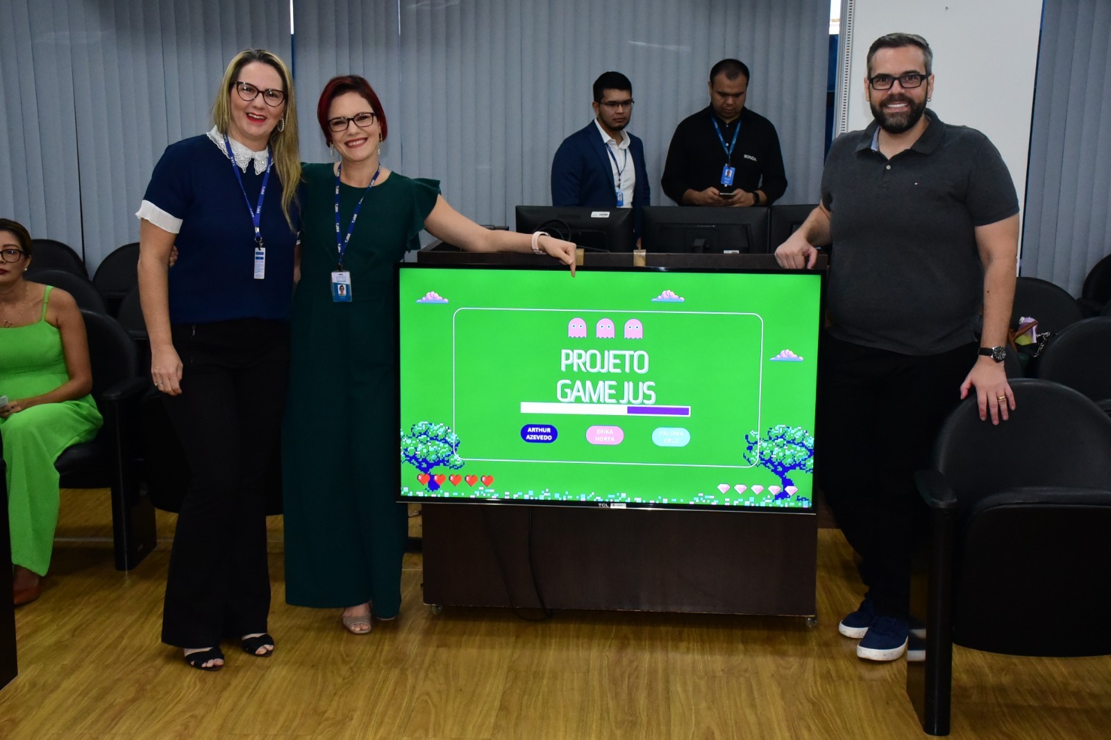 foto colorida com os duas servidoras mulheres e um homem ao lado do banner da TV no gamejus