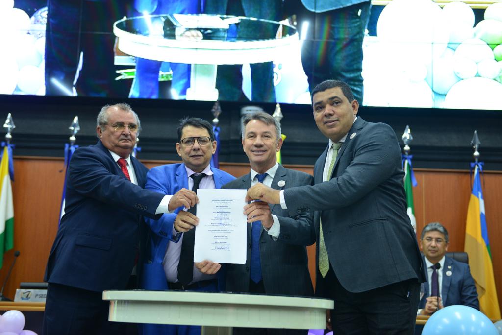 foto colorida mostrando o Presidente do TJRR, desembargador Jésus Nascimento no pulpito, ao lado está a mesa de autoridades compostas por representantes do Legislativo, Executivo e Judiciário. A frente da mesa contém  balões roxos, azul, amarelo e laranja em tons claros. 