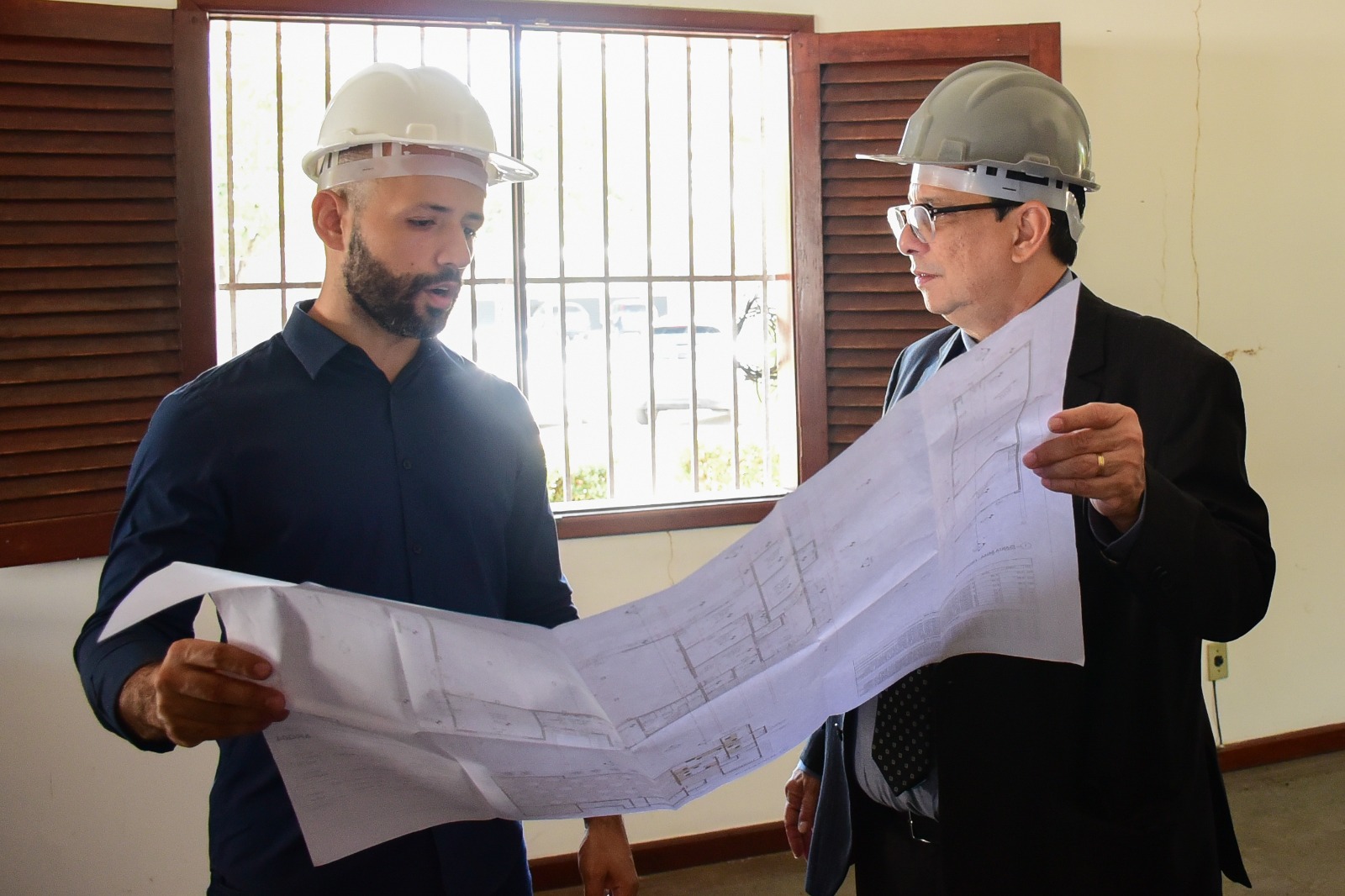 Foto colorida mostra o perfil do presidente do Poder Judiciário de Roraima, desembargador Jésus Nascimento  segurando uma planta, juntamente com o secretário de Infraestrutura e Logística do TJRR, Reubens Mariz, ambos usam capacetes. Ao fundo há uma janela de madeira gradeada, aberta.