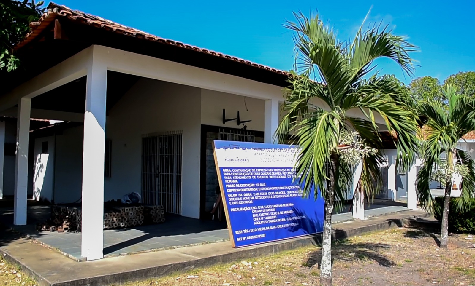 Foto colorida mostra uma estrutura com a  lateral arborizada. Encostada em uma coluna está uma com um placa azul com informações da contração. Abaixo o texto “INFRAESTRUTURA - Presidente do TJRR visita obras institucionais em andamento”.