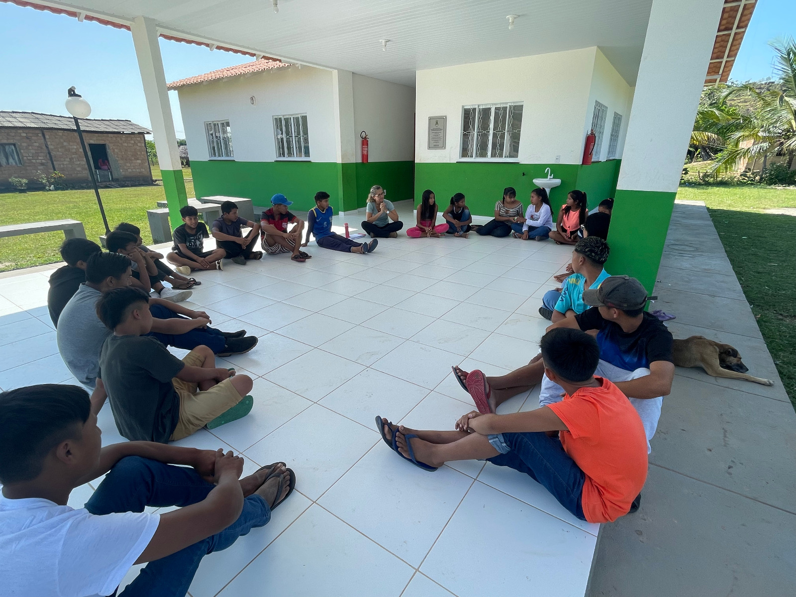 foto colorida na horizontal mostrando um pequeno grupo de comunidades sentadas no chão formando uma roda de conversa. Todos estão atentos às explicações de uma colaboradora da Coordenadoria de Violência Doméstica do Tribunal de Justiça de Roraima. 