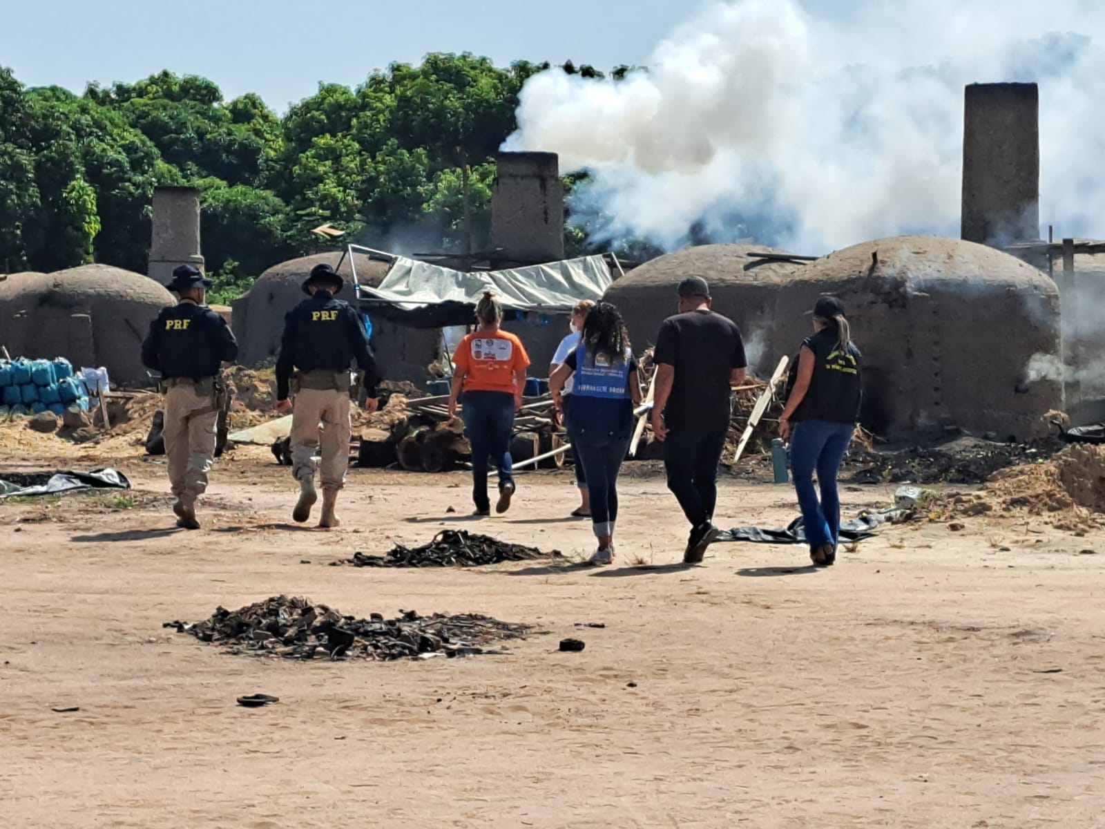 oto colorida mostra uma área aberta com três agentes da Polícia Rodoviária Federal, e quatro representantes do Trabalho de Erradicação do Trabalho Infantil, e do Centro de Referência Especializado de Assistência Social de Boa Vista, todos estão de costas para a câmera, caminhando em direção a diversas fornalhas de barro com fumaça saindo das chaminés. Ao redor do terreno há pequenos montes de lixo queimados e uma pequena uma área arborizada. 