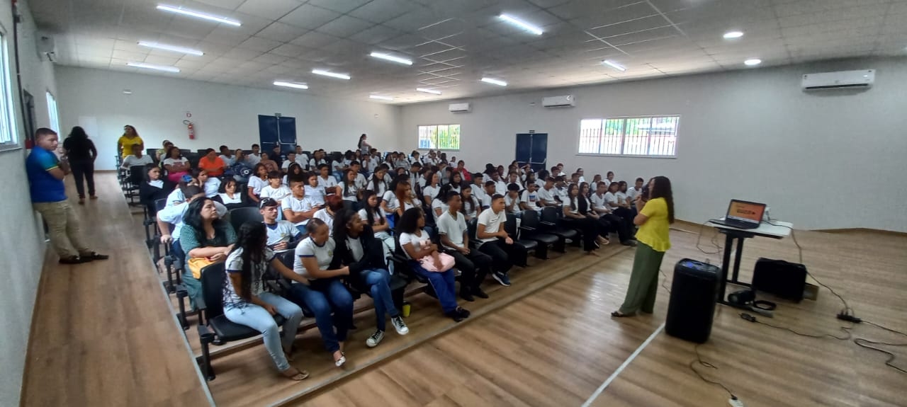 Foto colorida mostra um auditório com um  grupo de estudantes fardados, sentados em poltronas pretas, observando a palestrante. 