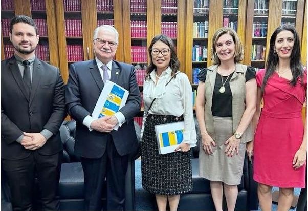  A foto colorida mostra o magistrado Cleber Gonçalves Filho, juíz de direito no Tribunal de Justiça de Roraima (TJRR), ao lado do ministro do Supremo Tribunal Federal (STF) Edson Fachin, e de mais três mulheres, posando para fotografia. No fundo da imagem contém uma estante de livros.