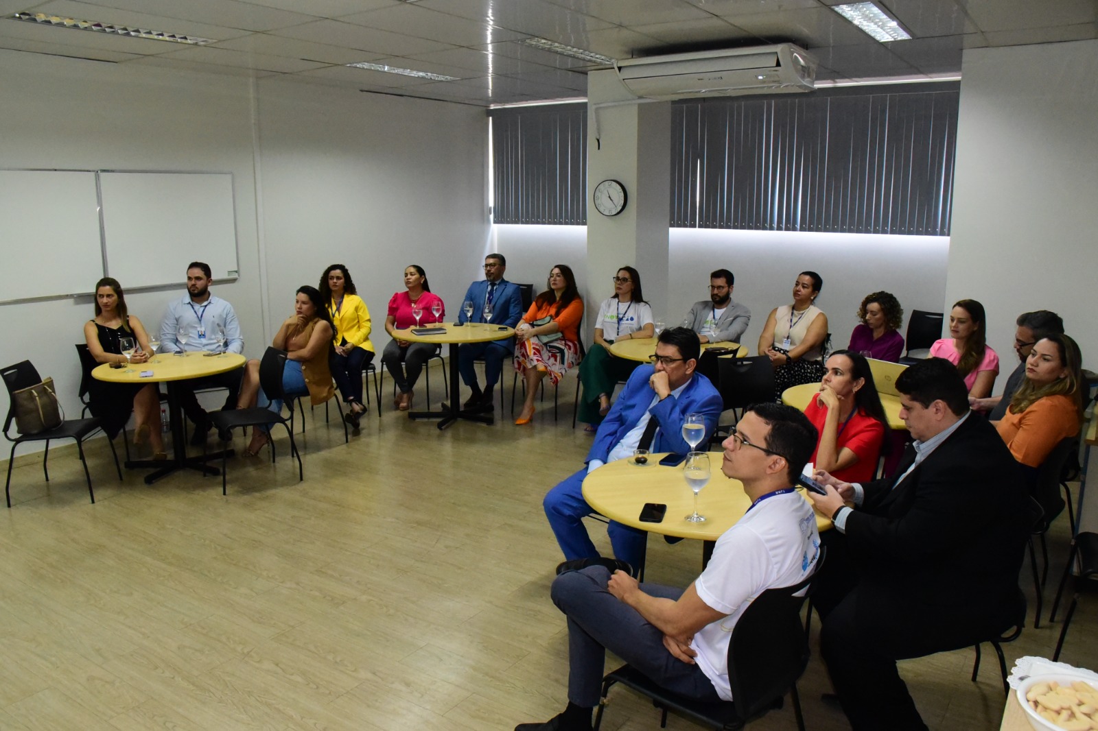 Imagem colorida com diversas pessoas, entre homens e mulheres, sentadas ao redor de mesas em uma sala de paredes brancas 