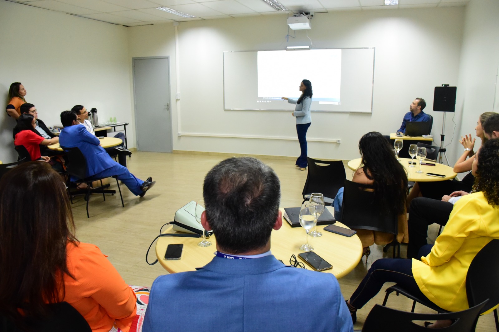 Imagem colorida mostra diversas pessoas sentadas em meia - lua ao redor de mesas enquanto assistem a apresentação de uma projeçâo que está sendo feita por uma mulher.